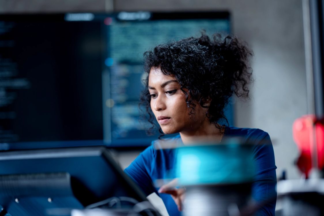 A woman with curly hair concentrating on a tablet in front of a monitor showing code.