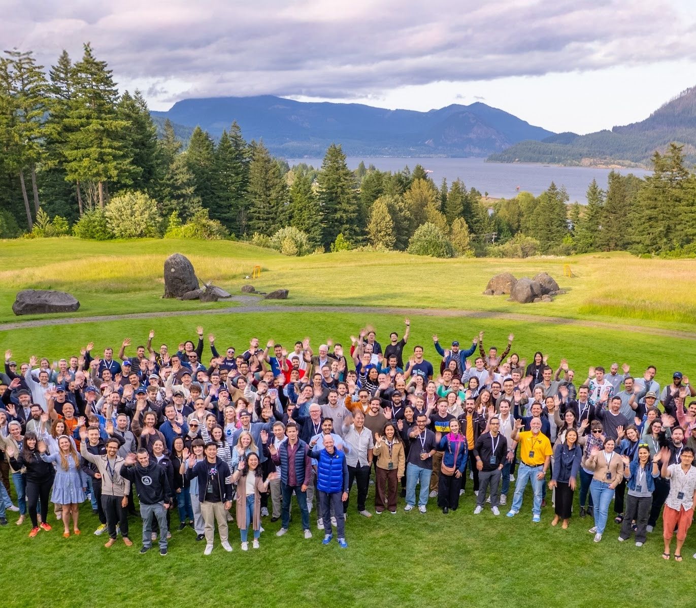 A large group of TRM employees stands together on a grassy lawn, smiling and waving at the camera, with evergreen trees, a river and mountains behind them.