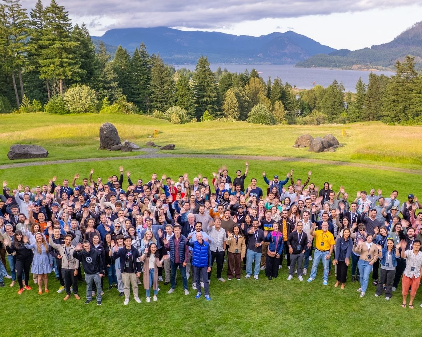 A large group of TRM employees stands together on a grassy lawn, smiling and waving at the camera, with evergreen trees, a river and mountains behind them.