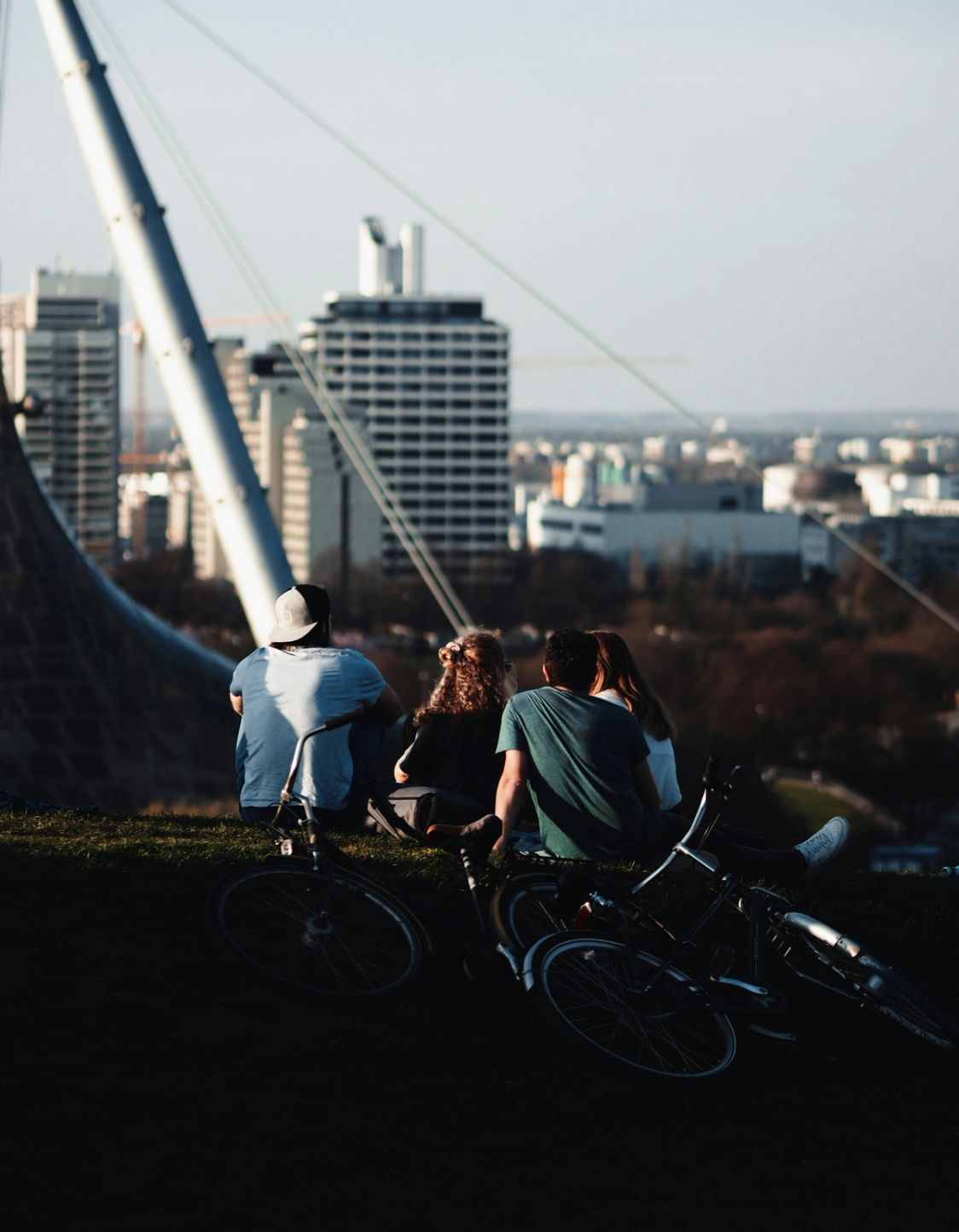 Three people sitting on a hillside near a bridge, looking out over a city skyline at dusk.