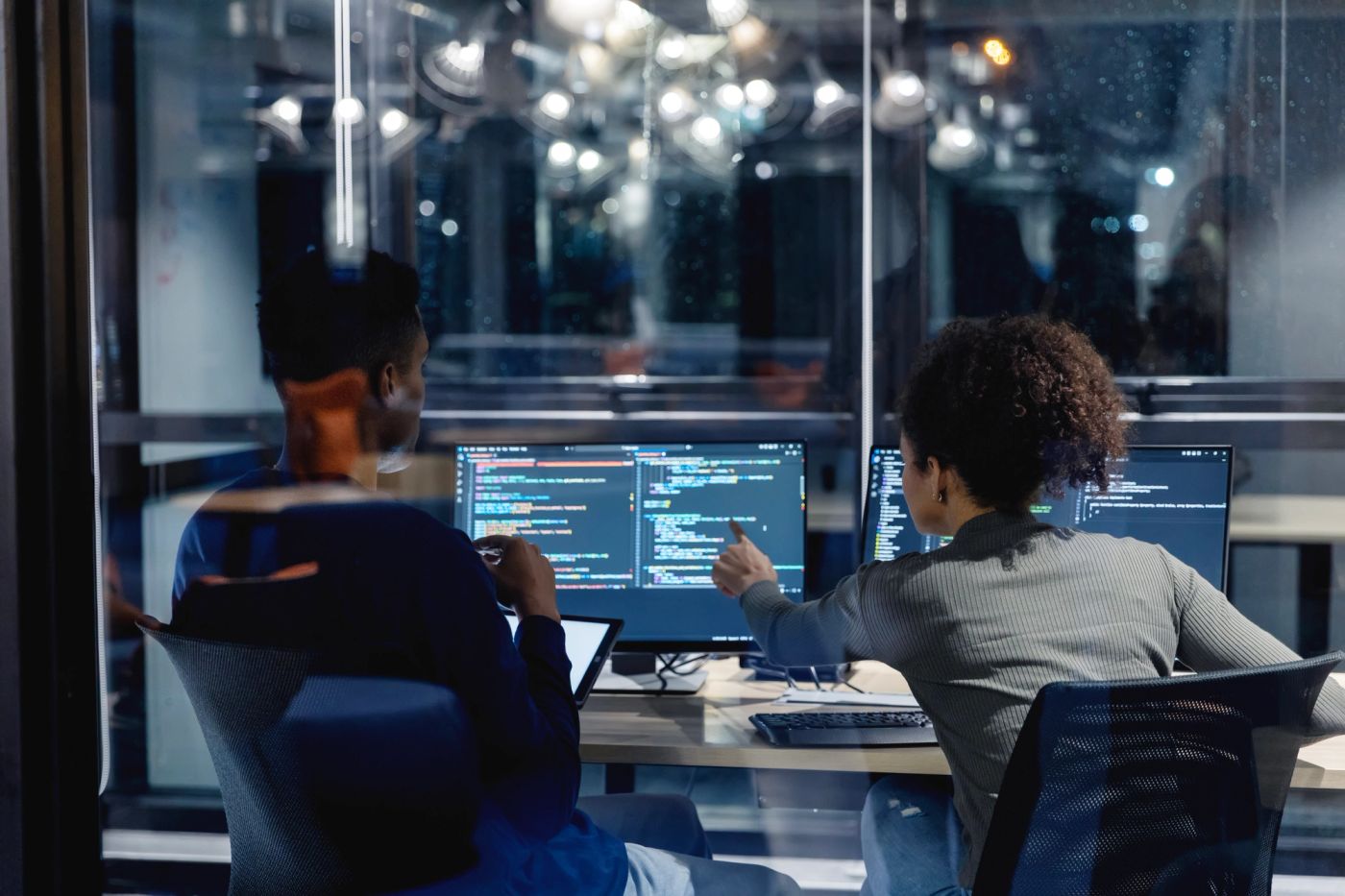 Two security analysts at desks in front of monitors showing code, seen through a glass wall.
