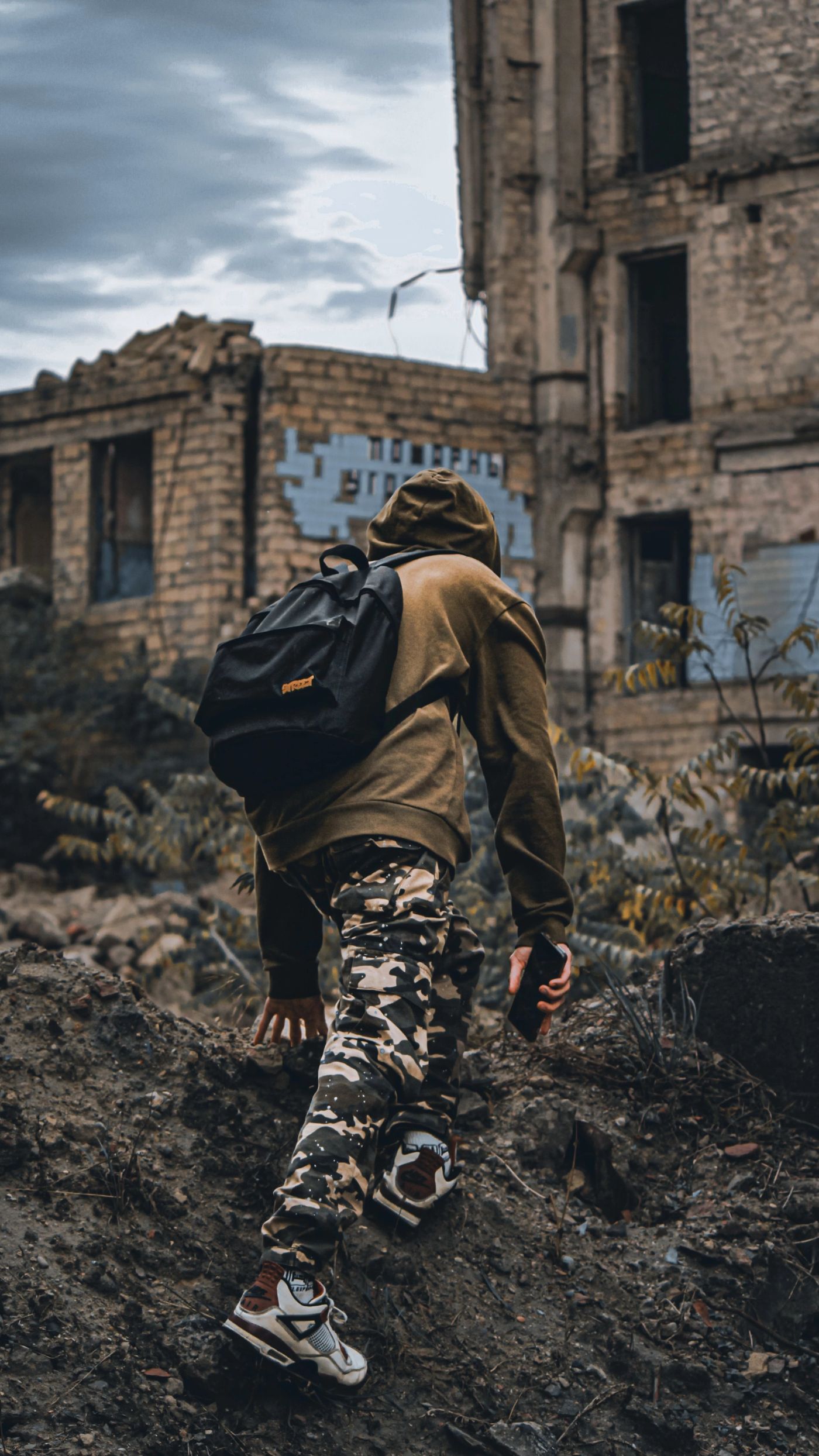 A person in camouflage clothing and a backpack walking uphill past a ruined, graffitied building.