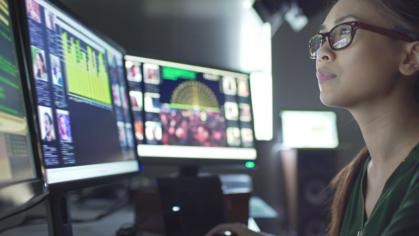 A woman with glasses looking at a wall of monitors showing profiles and a map.