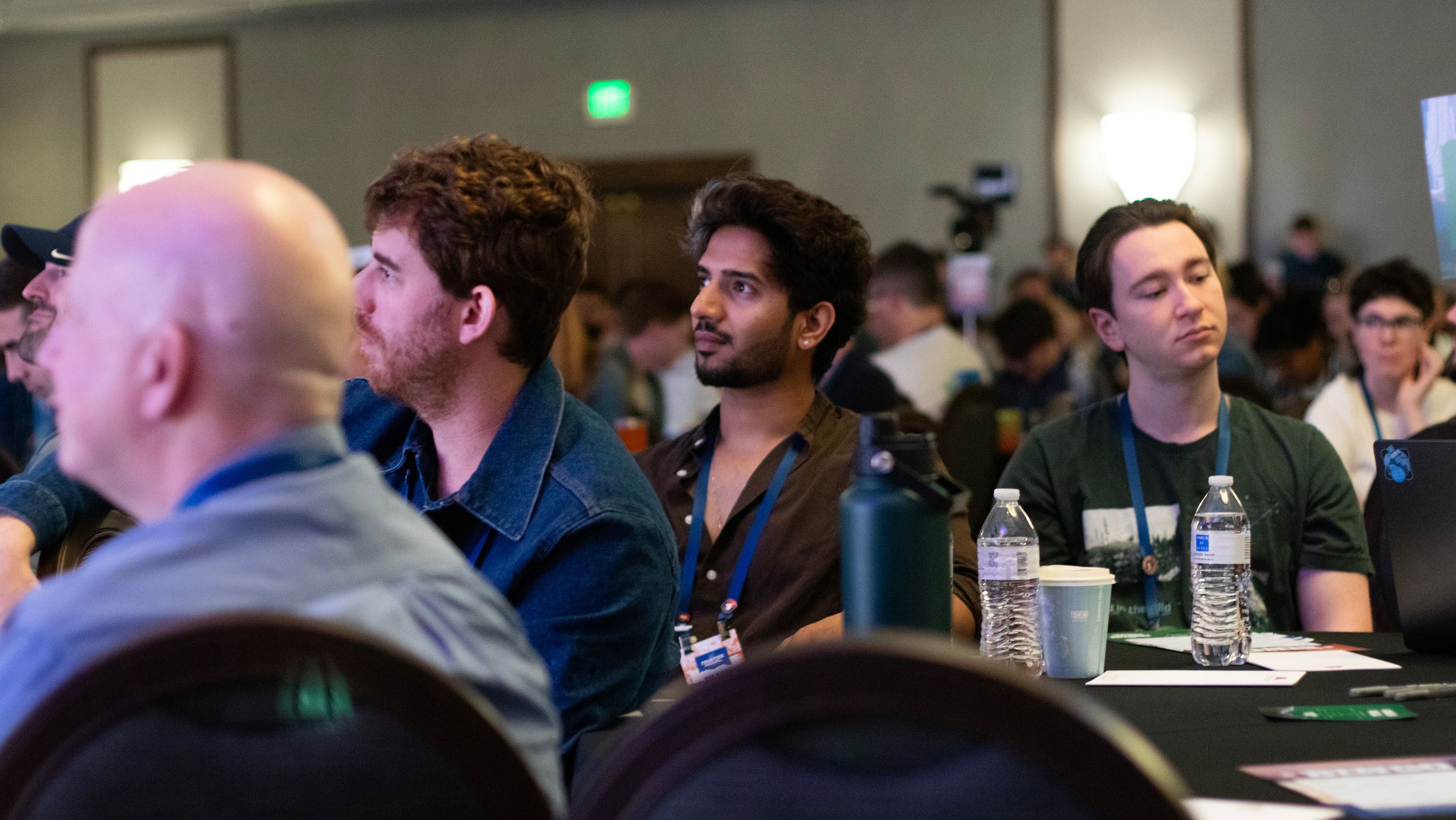 Attendees in a conference room listening to a speaker; two men in the front rows look on.