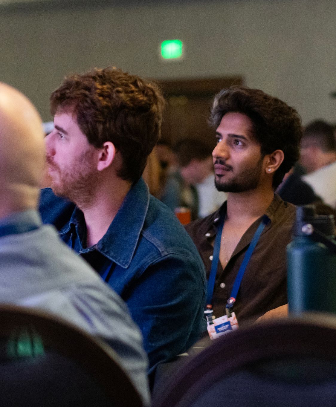 Attendees in a conference room listening to a speaker; two men in the front rows look on.