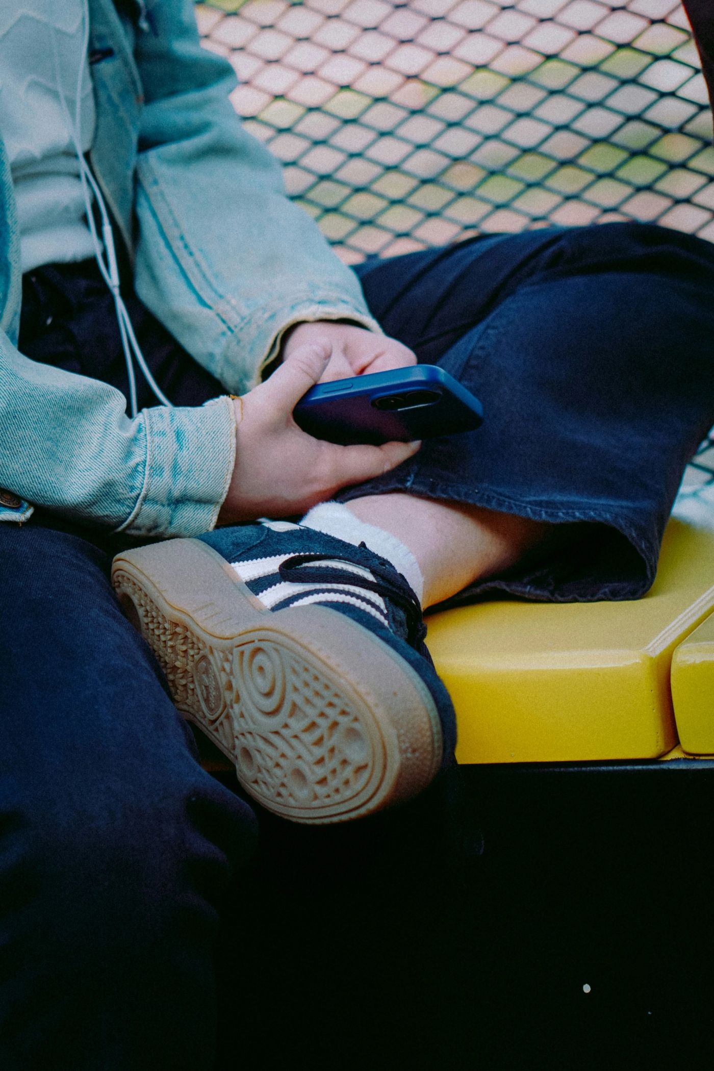 A seated person in a denim jacket and wide trousers holding a smartphone, on a yellow bench.