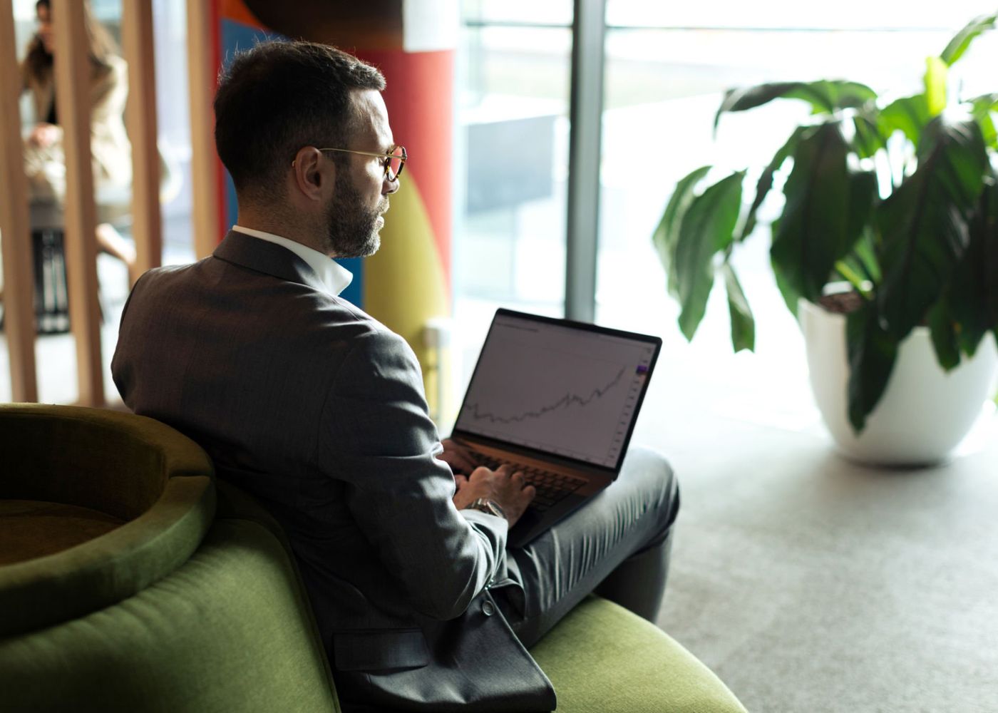 A man in a suit and glasses sitting on a green sofa, working on a laptop that shows a line chart.
