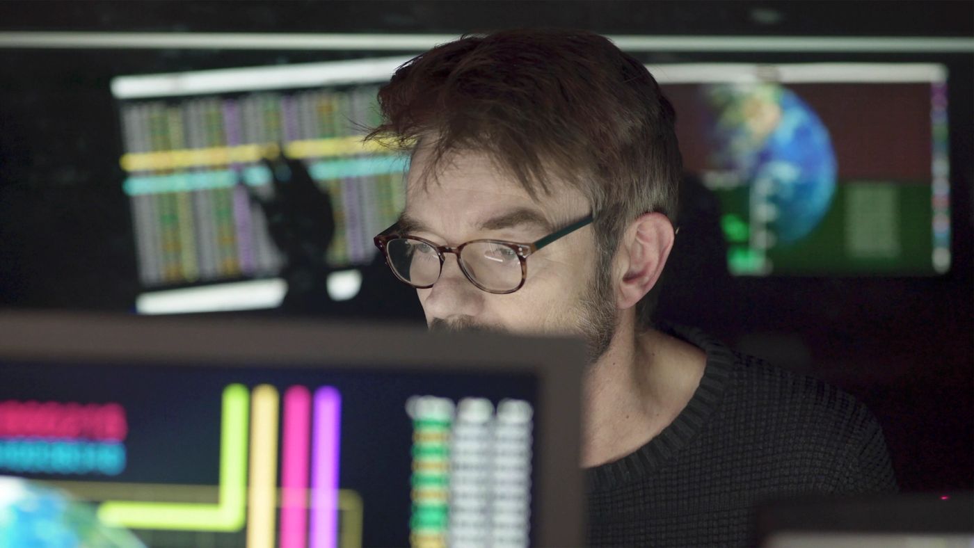 A man with glasses studying monitors that show trading data and a globe.