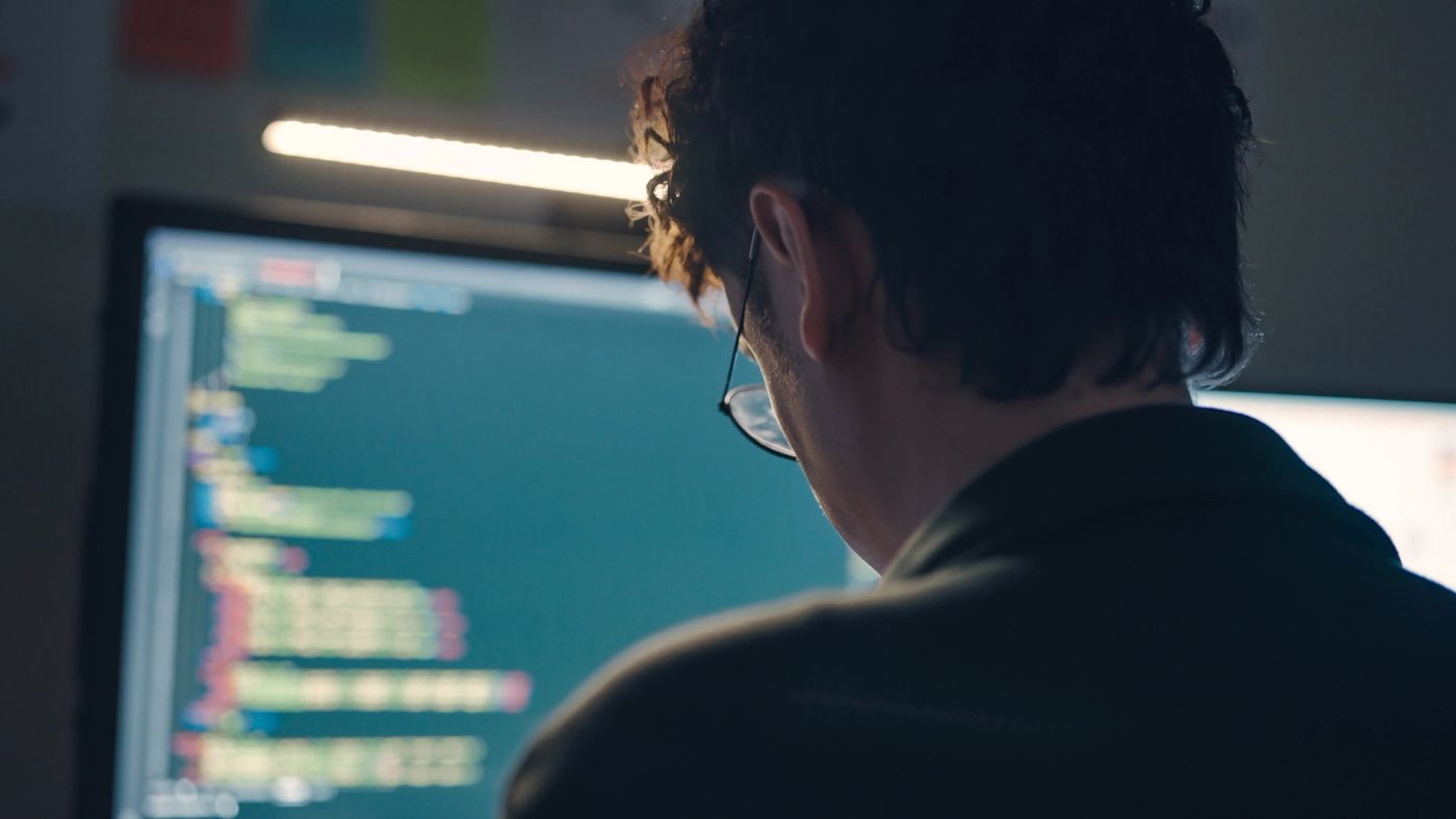 A software engineer seen from behind, looking at code on a monitor.