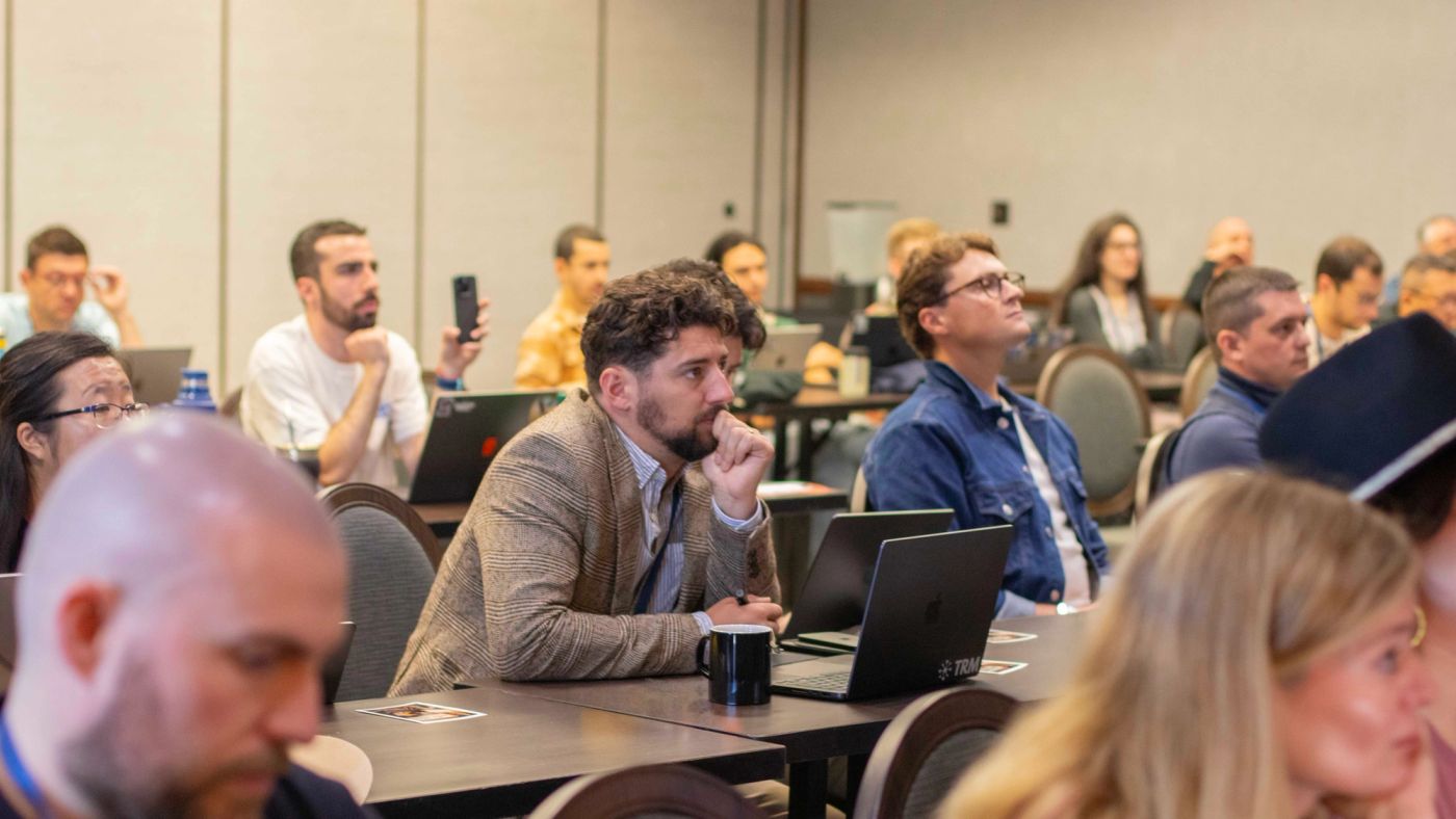 Attendees seated in a conference room, watching a presentation; a man in the centre rests his chin on his hand.