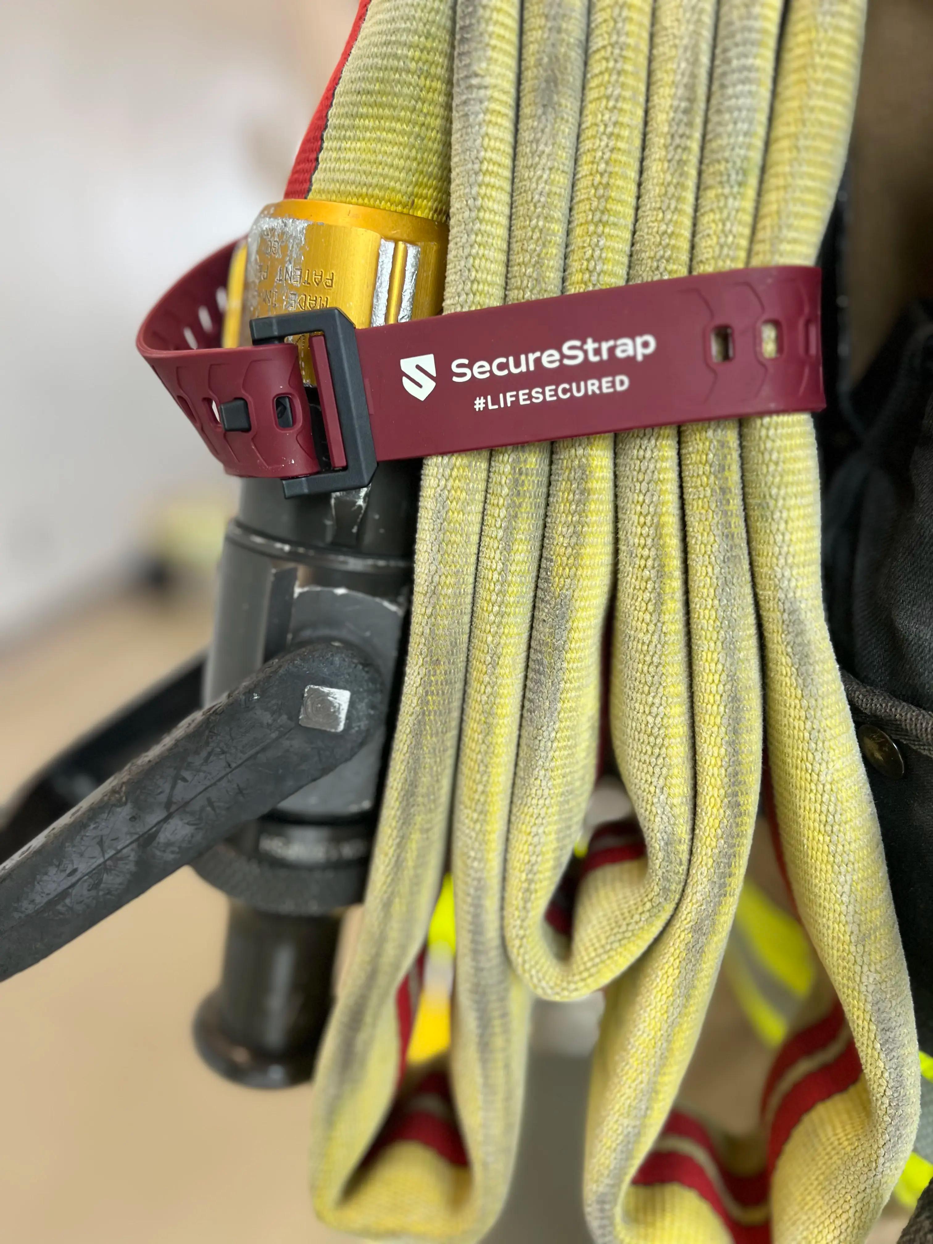 Close-up of a folded yellow fire hose secured with a red SecureStrap labeled “SecureStrap #LIFESECURED.” A metal valve or nozzle with a black handle is attached to the hose. The image focuses on the strap and hose texture.