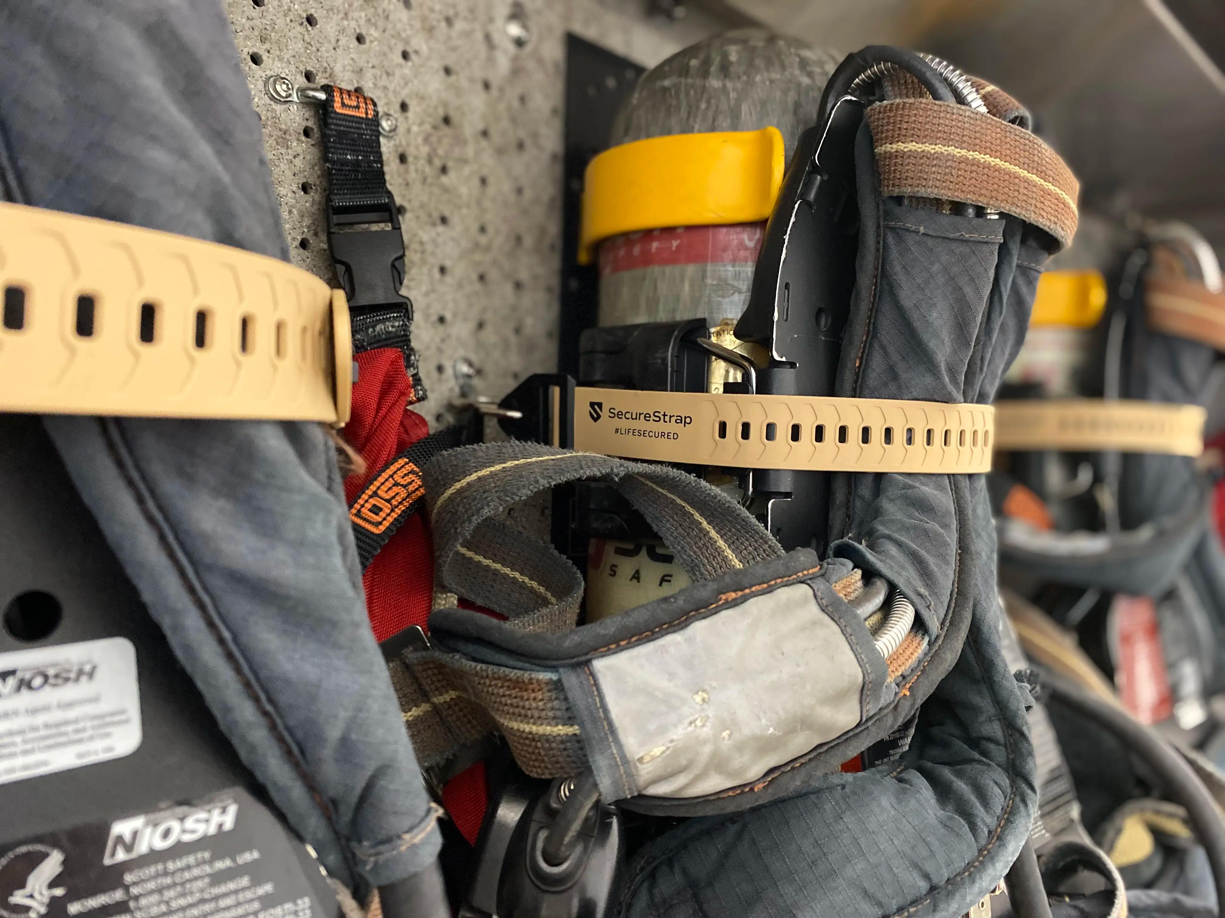 Close-up of firefighter breathing apparatuses mounted on a wall. Each unit is secured with a tan SecureStrap labeled “SecureStrap #LIFESECURED.” The gear includes air tanks, harnesses, and straps, with visible NIOSH certification labels.