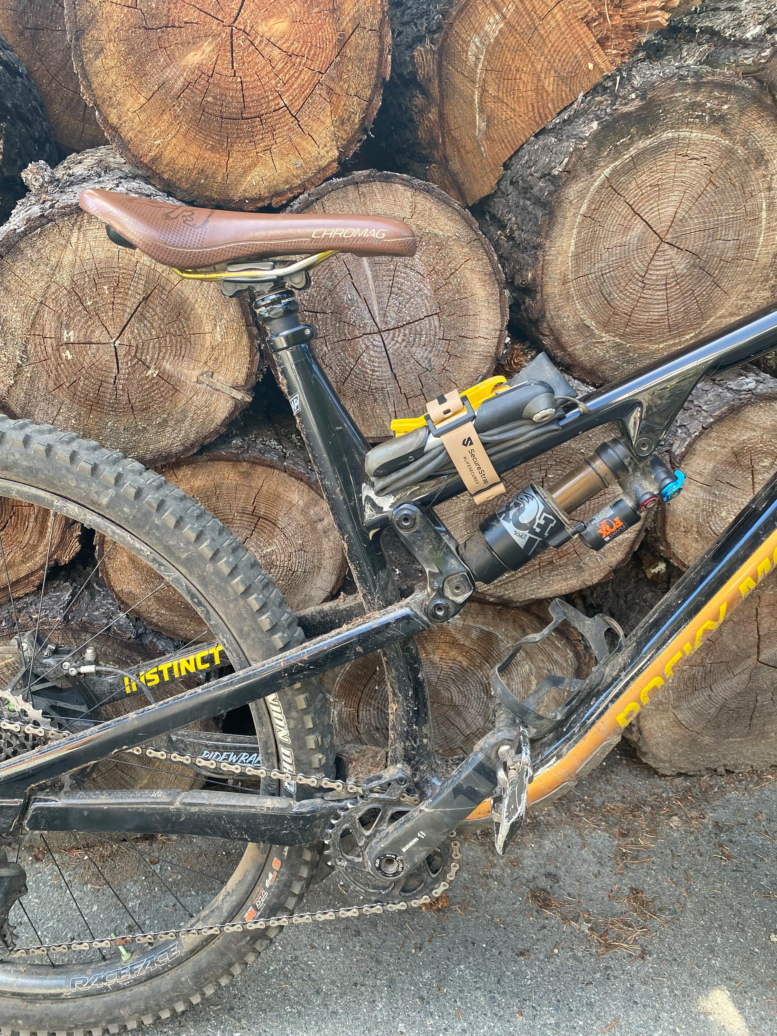 A black and orange Rocky Mountain Instinct mountain bike is parked in front of a stack of cut logs. A beige SecureStrap holds a repair kit and tools to the bike frame under the seat. The bike has a brown Chromag saddle and visible dirt on the frame and tires.
