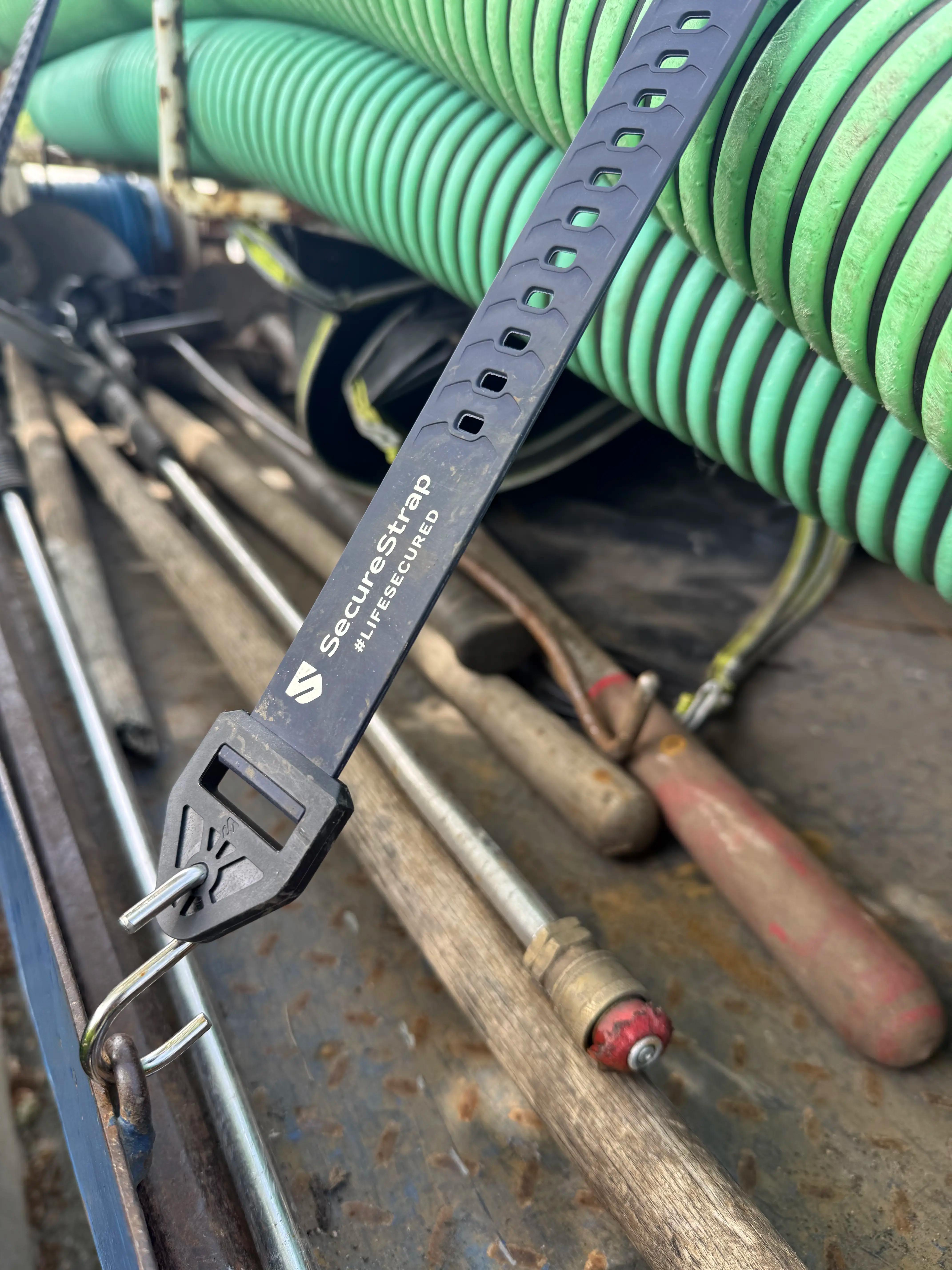 A black SecureStrap with the label “SecureStrap #LIFESECURED” is hooked onto a metal loop. In the background, coiled green hoses and several long-handled tools with wooden and metal handles are scattered on a rusty, textured metal surface.