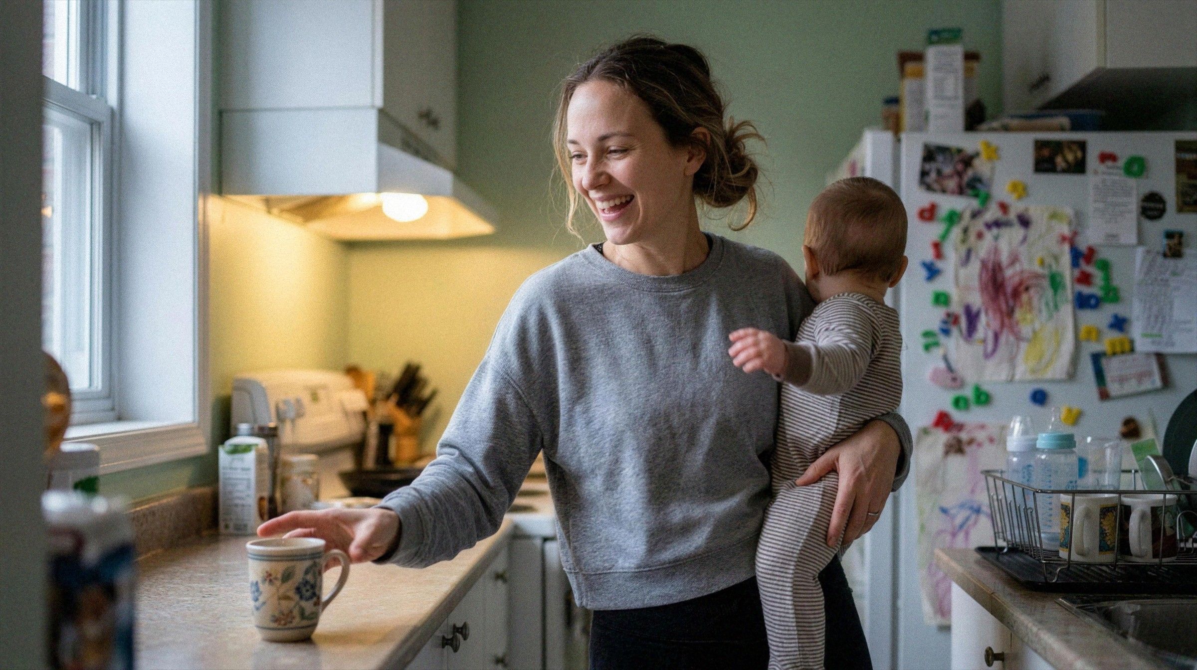 A mother in her kitchen laughs while holding her toddler on her hip in soft morning light.
