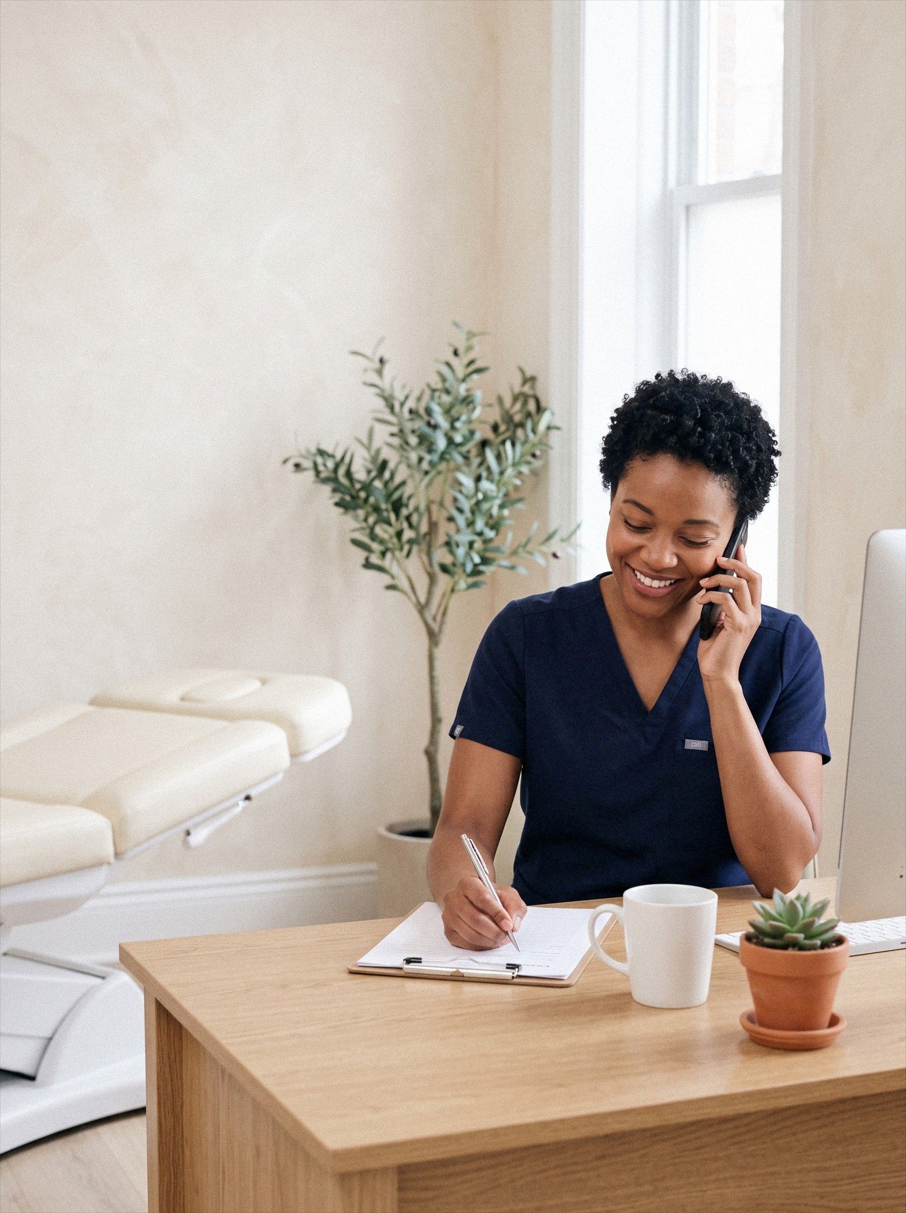 A nurse at Ideal Body Clinic takes a patient's call at her desk and writes notes on a clipboard.