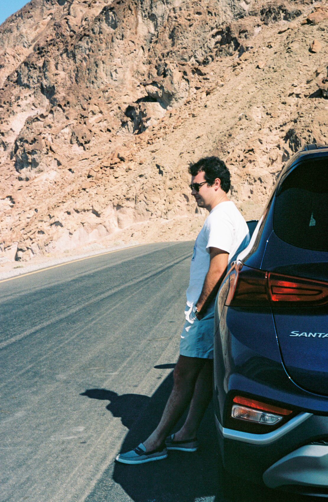 A man wearing a white t-shirt and sunglasses leans on a car on the side of the road in Death Valley.