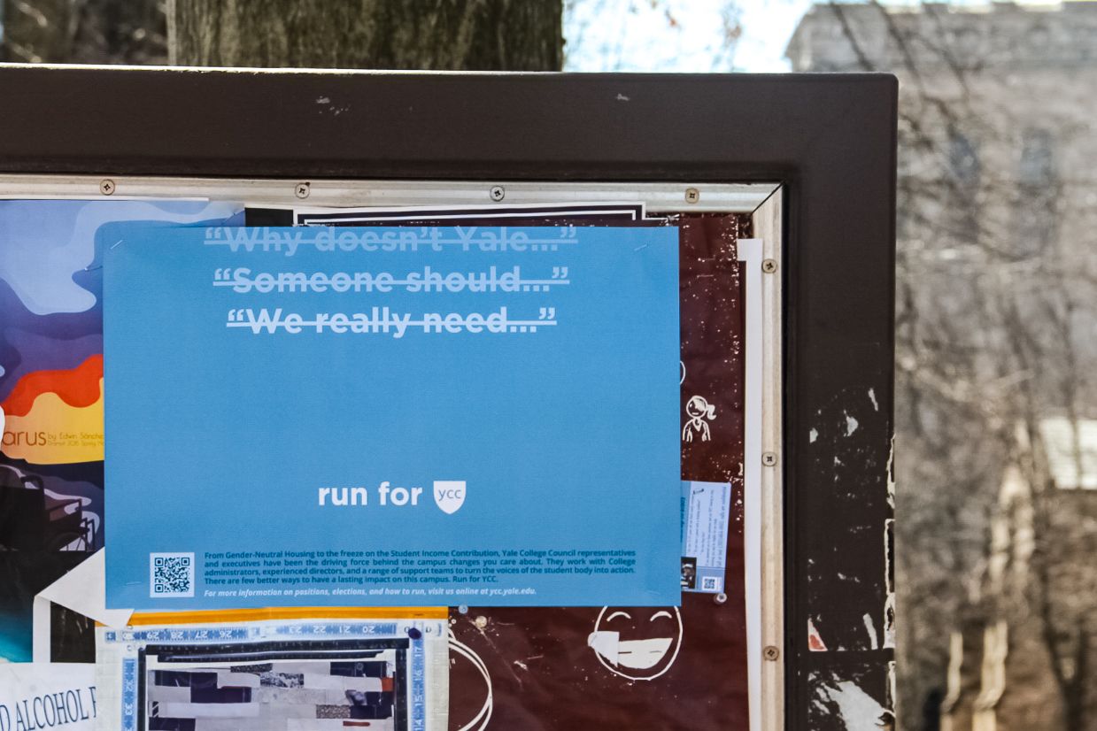 A blue poster on a bulletin board. At the top of the poster are three lines of text with lines strikethrough lines: Why doesn’t Yale... Someone should... We really need... and below it, the text: run for YCC