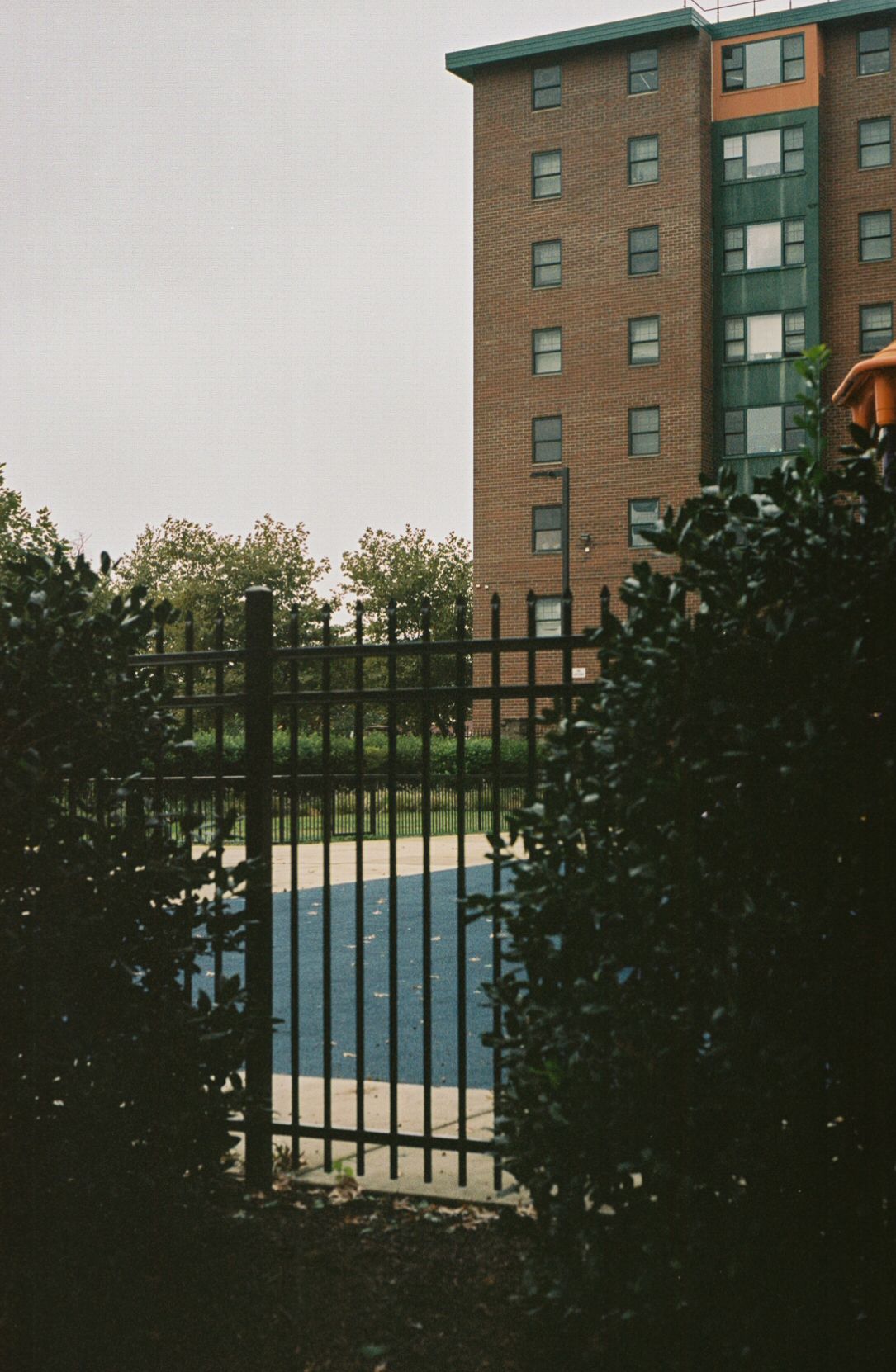 A 35mm film street photograph of a brick apartment building in Washington, DC. The apartment building has green and orange details. A blue courtyard is visible through a steel fence.
