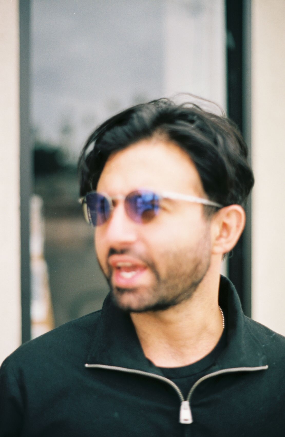 An out-of-focus portrait of a young man. He has long black hair and a short beard, and is wearing purple sunglasses and a black sweater.