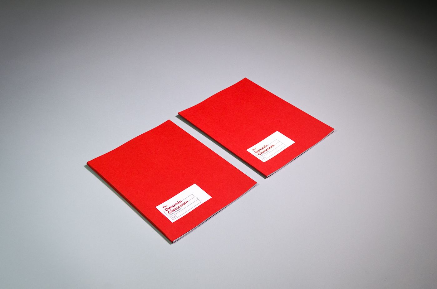 Two copies of the book. It measures about 8 inches wide by 10.5 inches tall. The cover is bright red matte paper. A small white label is adhered towards the bottom left corner. On it is printed a small black table of information, with “The Dynamic Classroom” overprinted in large red letters.