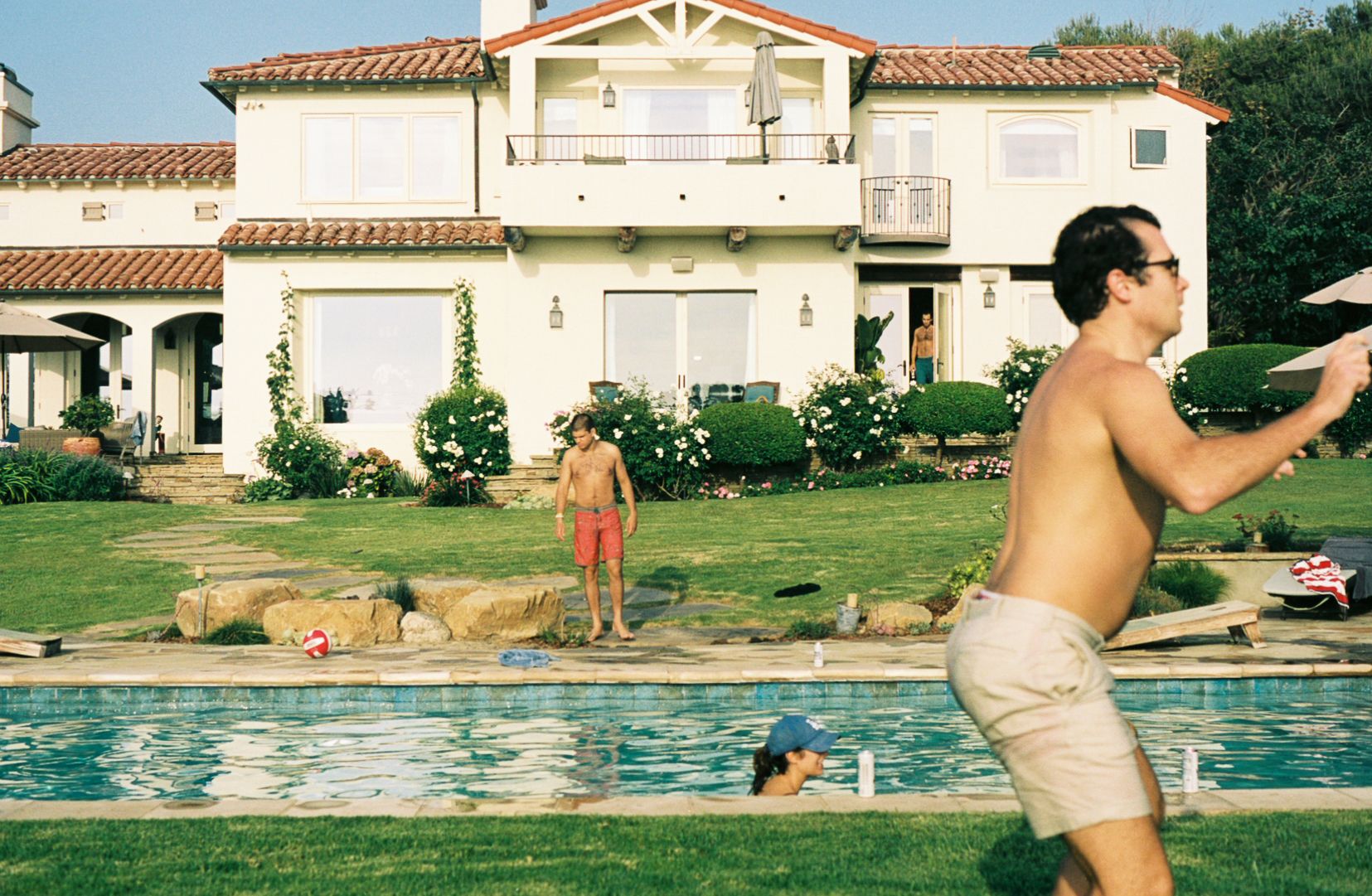 A view of the back of a house with a tile roof in California. In the foreground, a woman swims in a pool and a man catches a frisbee.