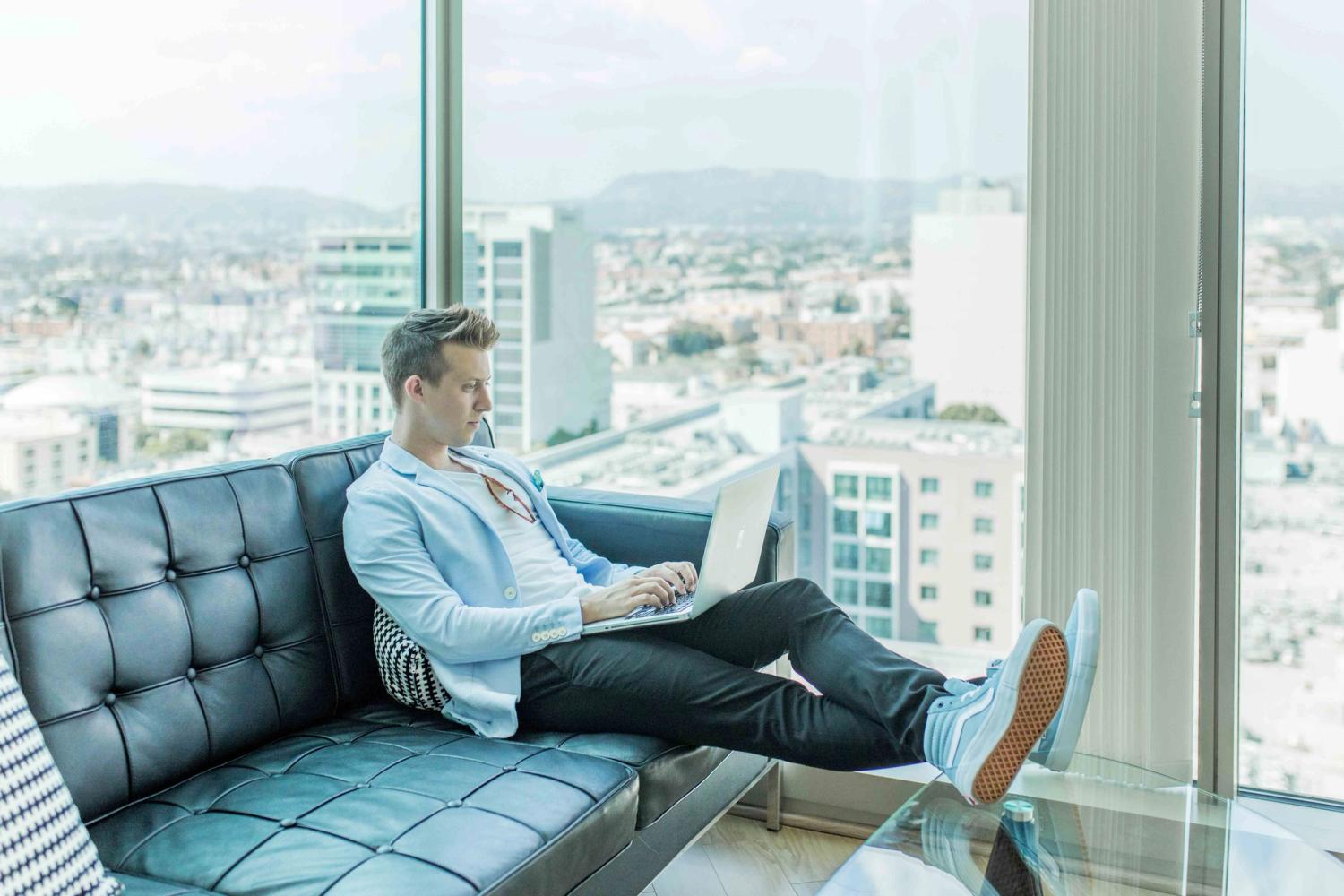 A young man laying on the sofa while working on his laptop with a city view behind him