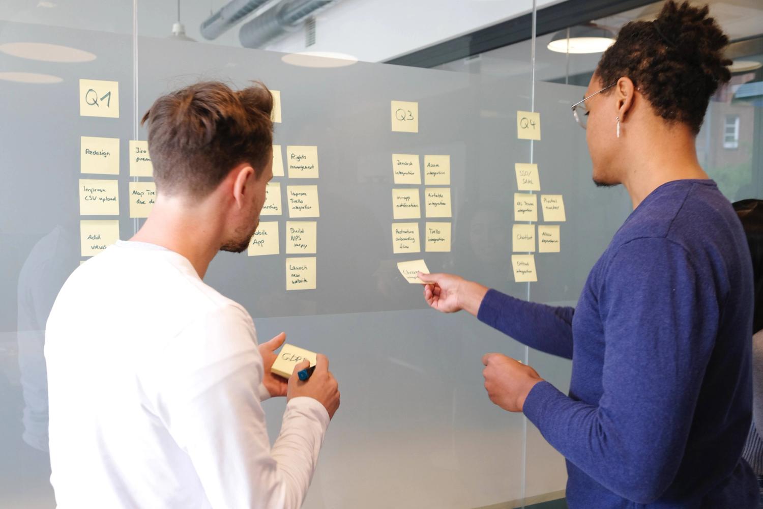 One white male with white t-shirt and one black male with blue shirt are placing sticky notes on a wall to create a strategy