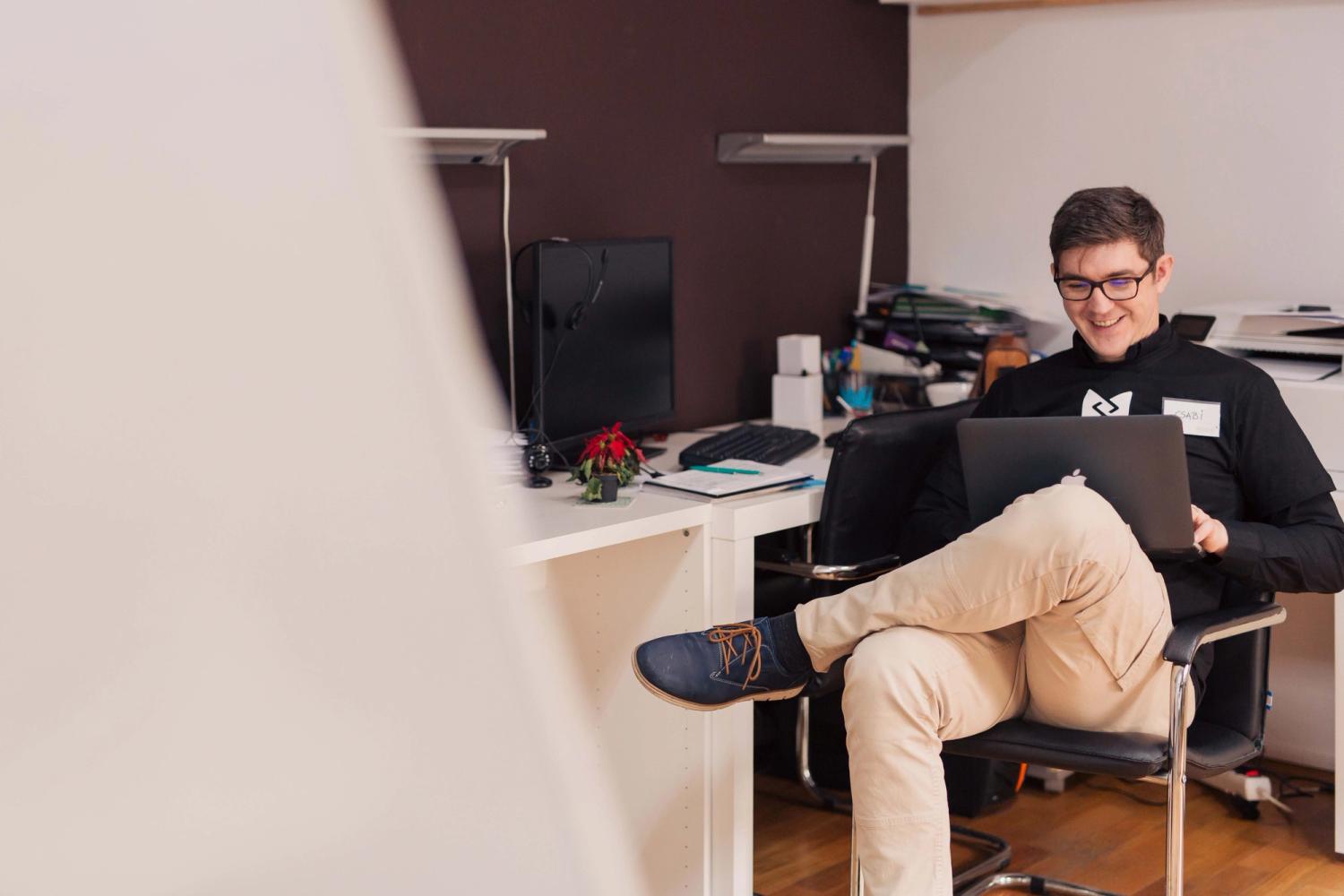 A guy sits in a chair while working on his laptop in his office smiling at the screen
