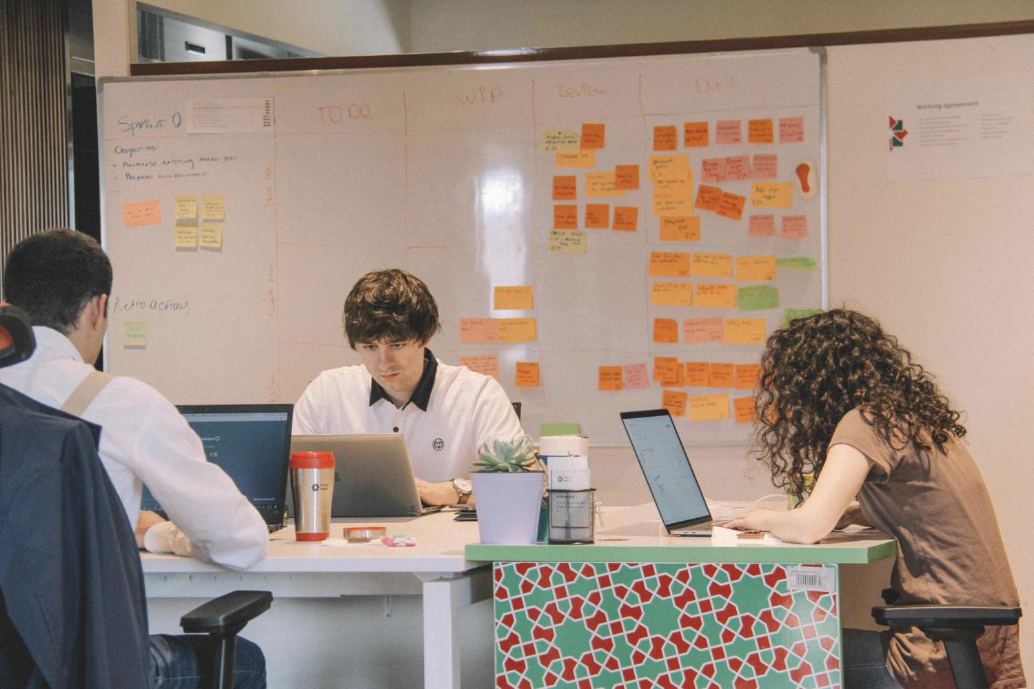 three people working on their agile development while having a white board with sticky notes and different drawings in the background for their agile startup