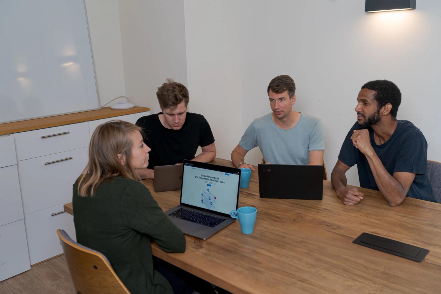 four collagues speaking about their work while sitting on chairs next to a wooden table with open laptops on it
