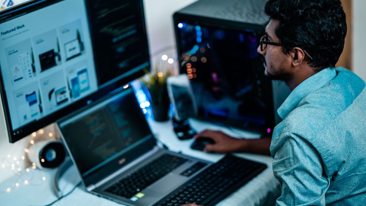 South Asian student working at a computer setup with laptop and monitor, studying computer science and software development
