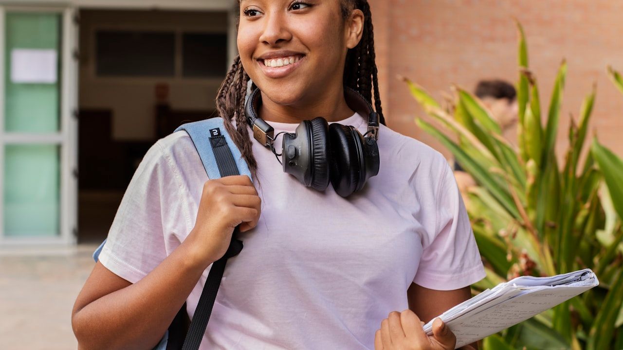 A smiling student with a backpack, headphones around her neck and notebooks in hand, standing outside a school building