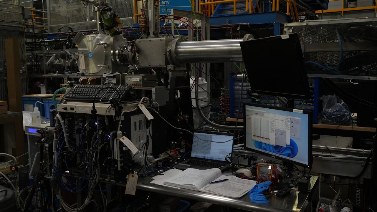 Engineering laboratory workstation with complex scientific machinery, computer monitors displaying data, and open technical documents