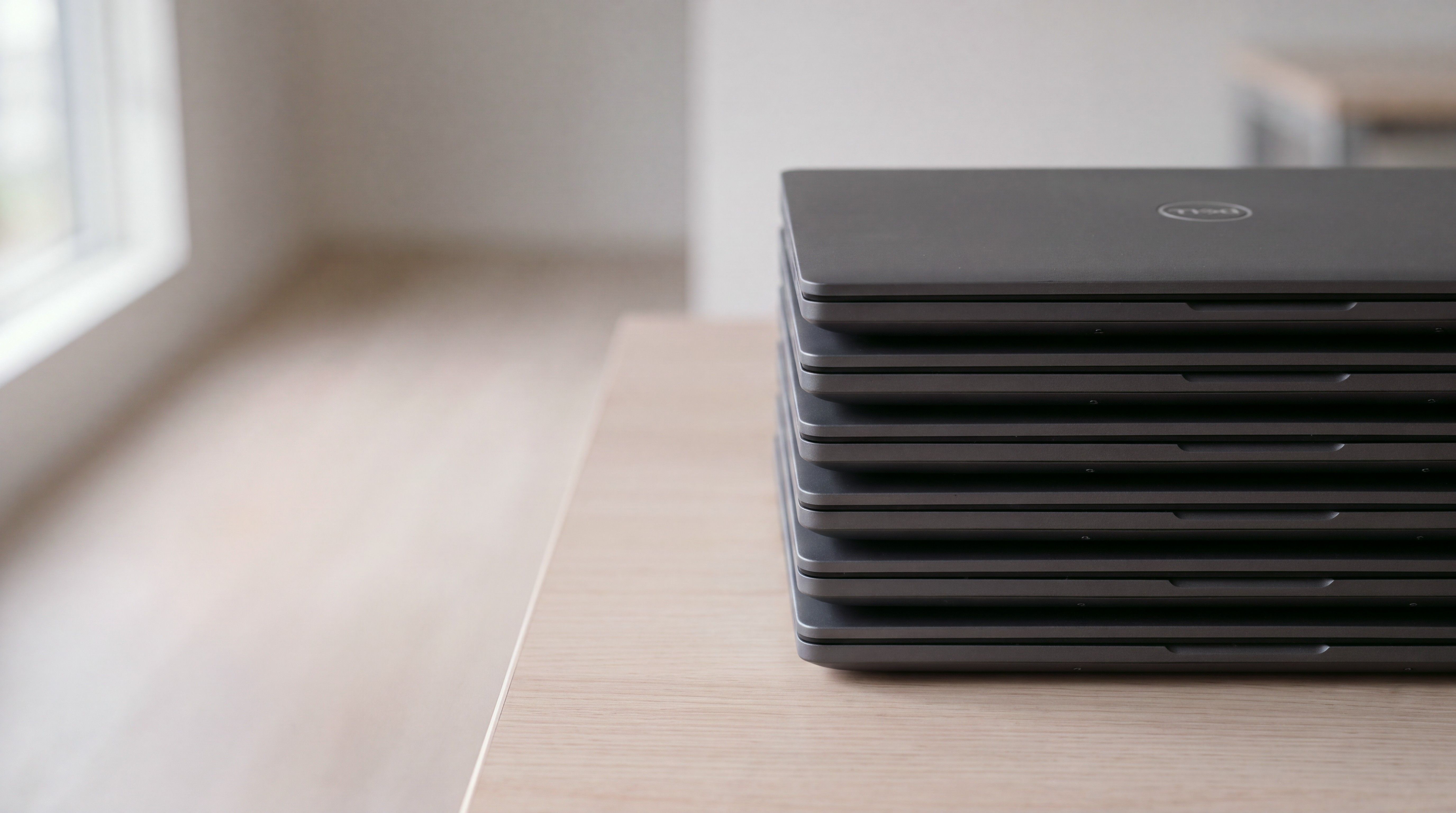 Six identical closed Dell laptops stacked neatly on a pale wood table.