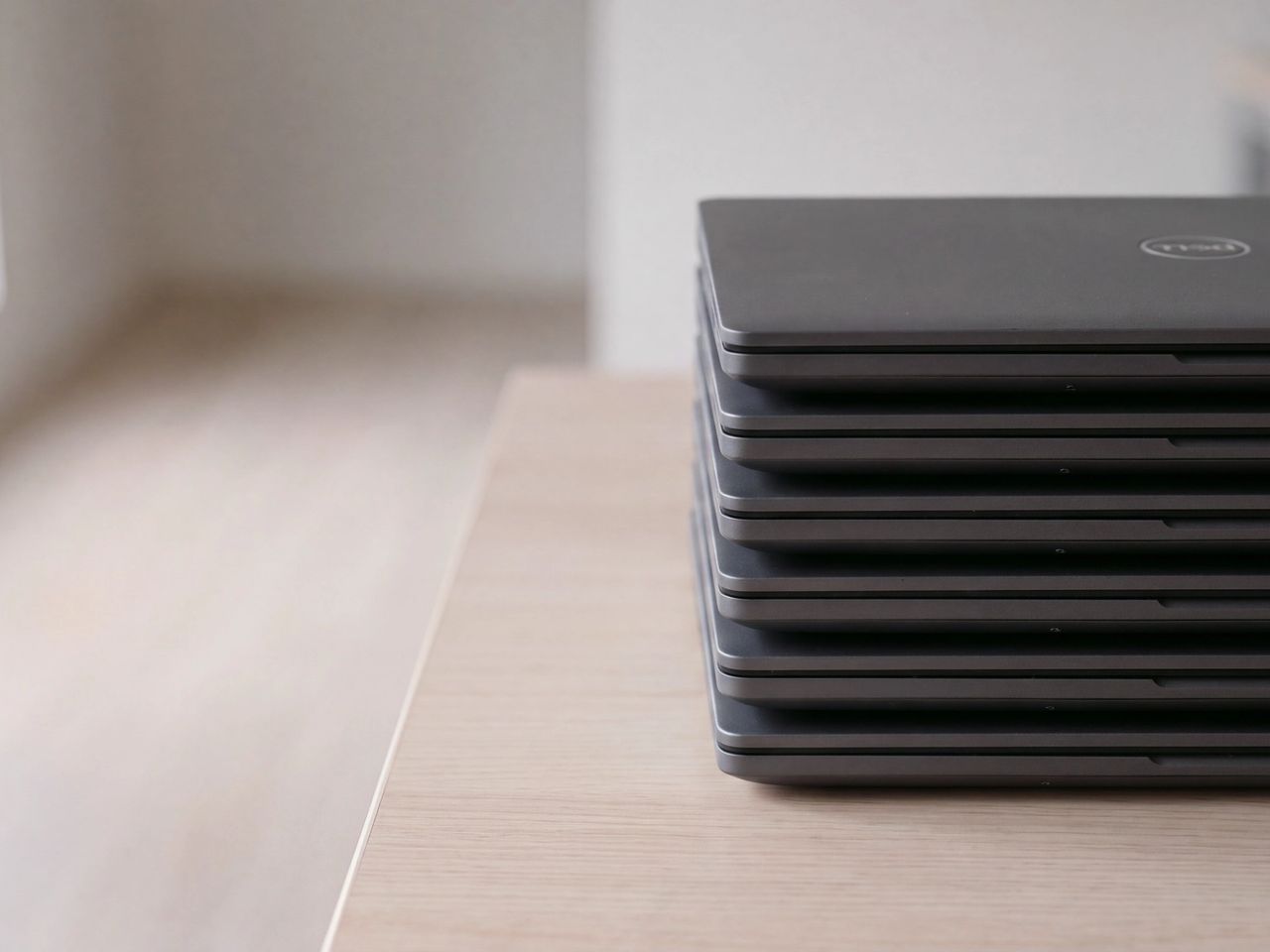 Six identical closed Dell laptops stacked neatly on a pale wood table.