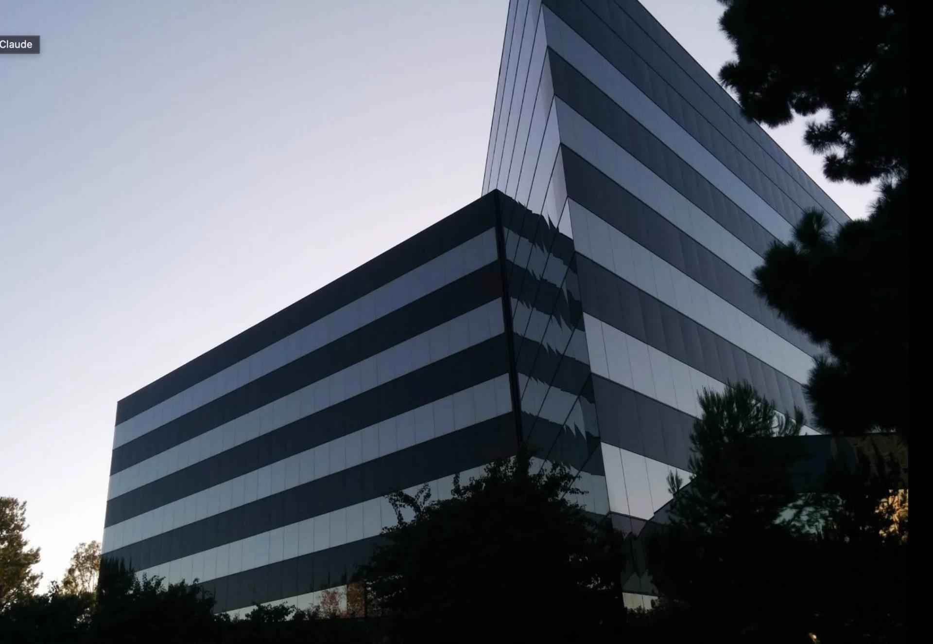 The s90 office building at 4590 MacArthur Blvd, Newport Beach: a dark glass mid-rise photographed from below at dusk, horizontal banding across the facade, tree branches across the upper corner.