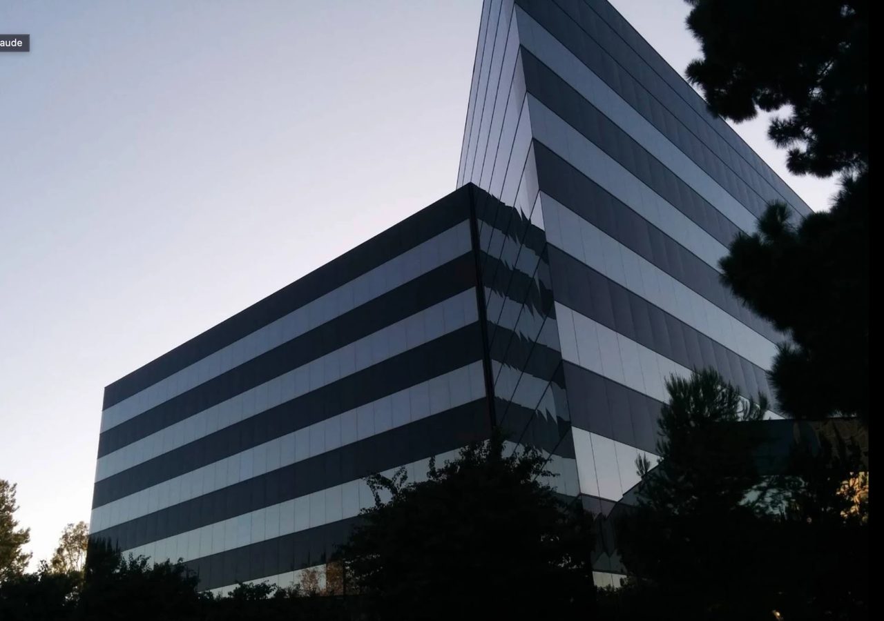 The s90 office building at 4590 MacArthur Blvd, Newport Beach: a dark glass mid-rise photographed from below at dusk, horizontal banding across the facade, tree branches across the upper corner.
