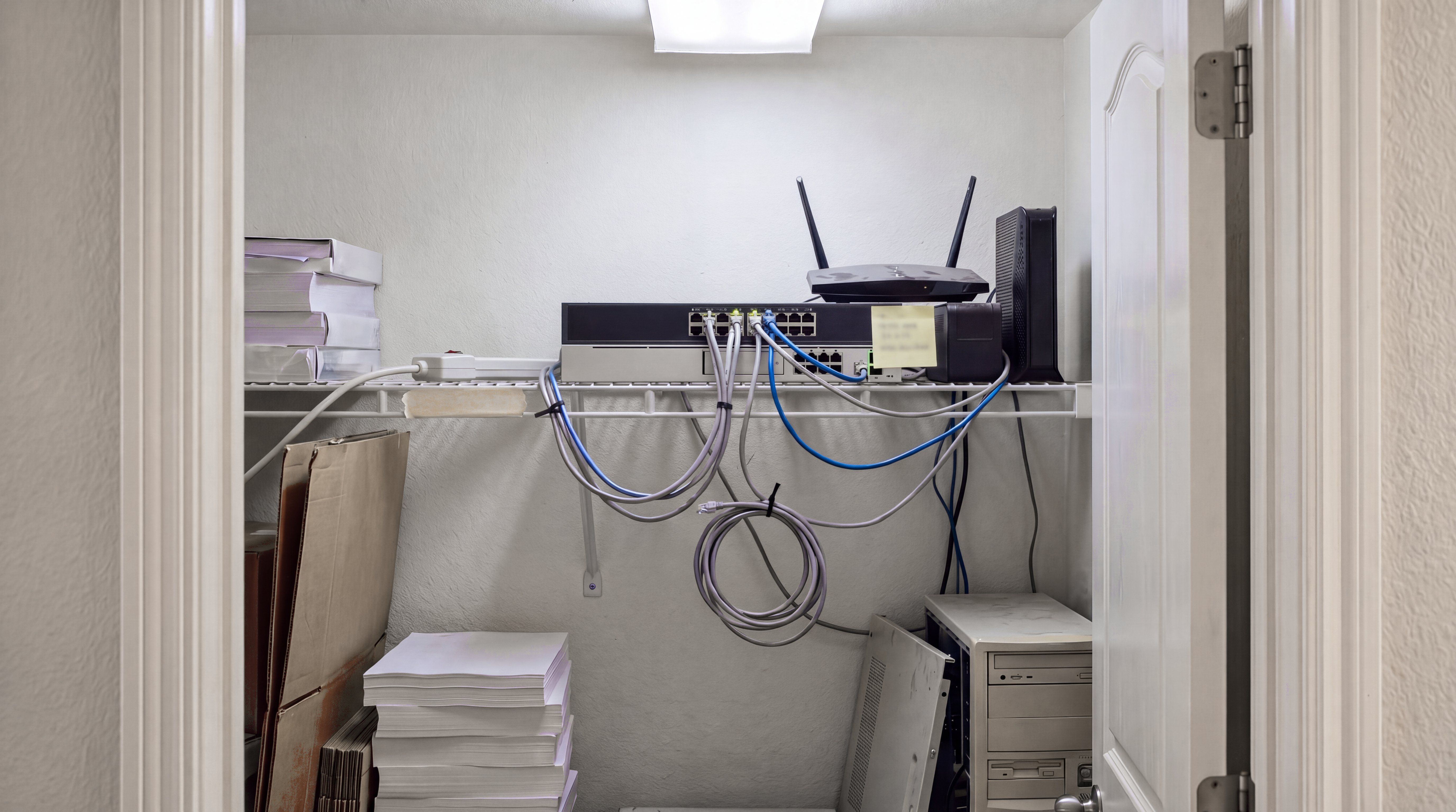 A small office supply closet where a network switch, router and modem sit stacked on a wire shelf with loose cables, beside stored paper and an old desktop tower.
