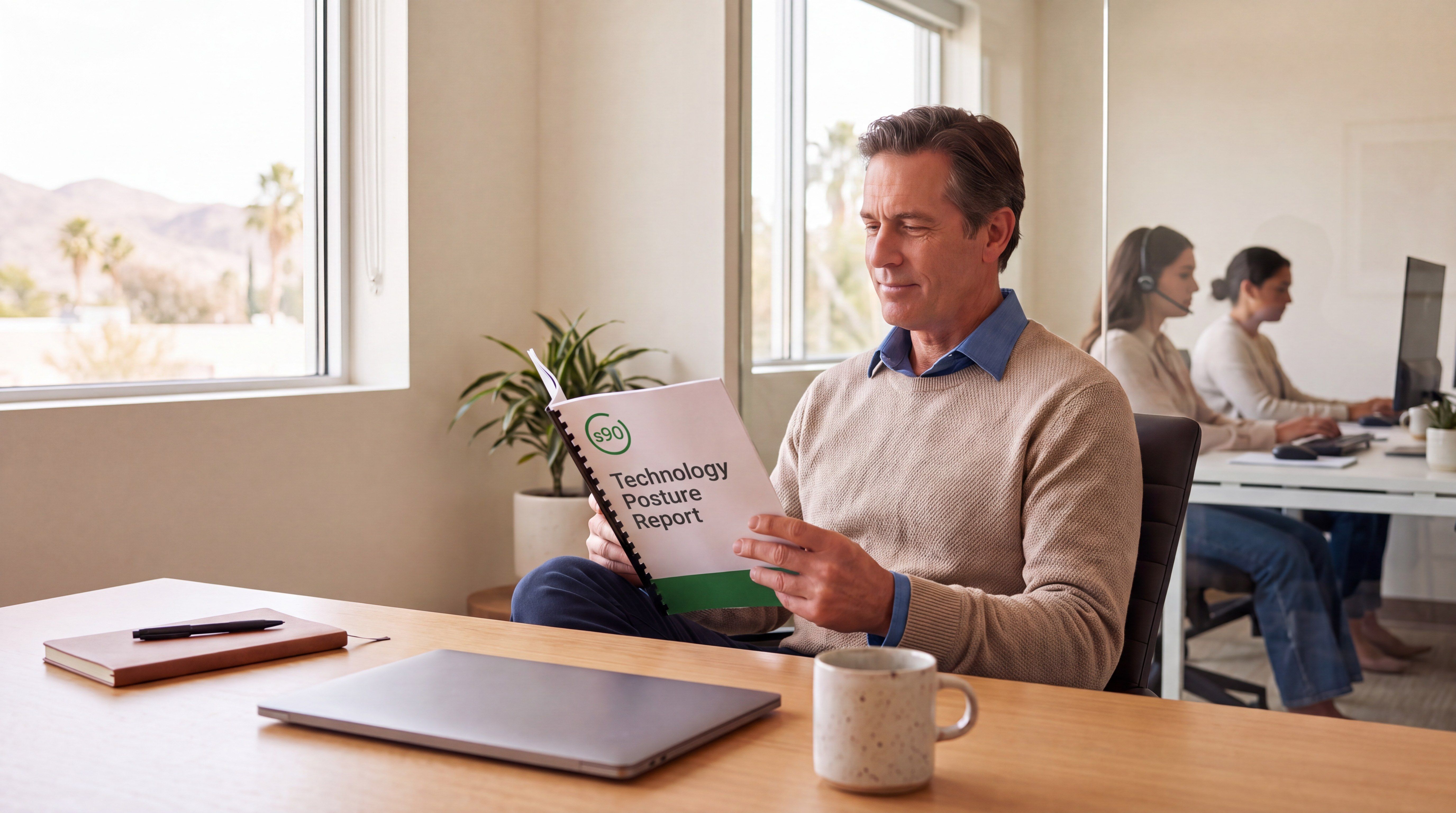 A business owner reads a bound quarterly report at their desk while staff work quietly in the office behind them.