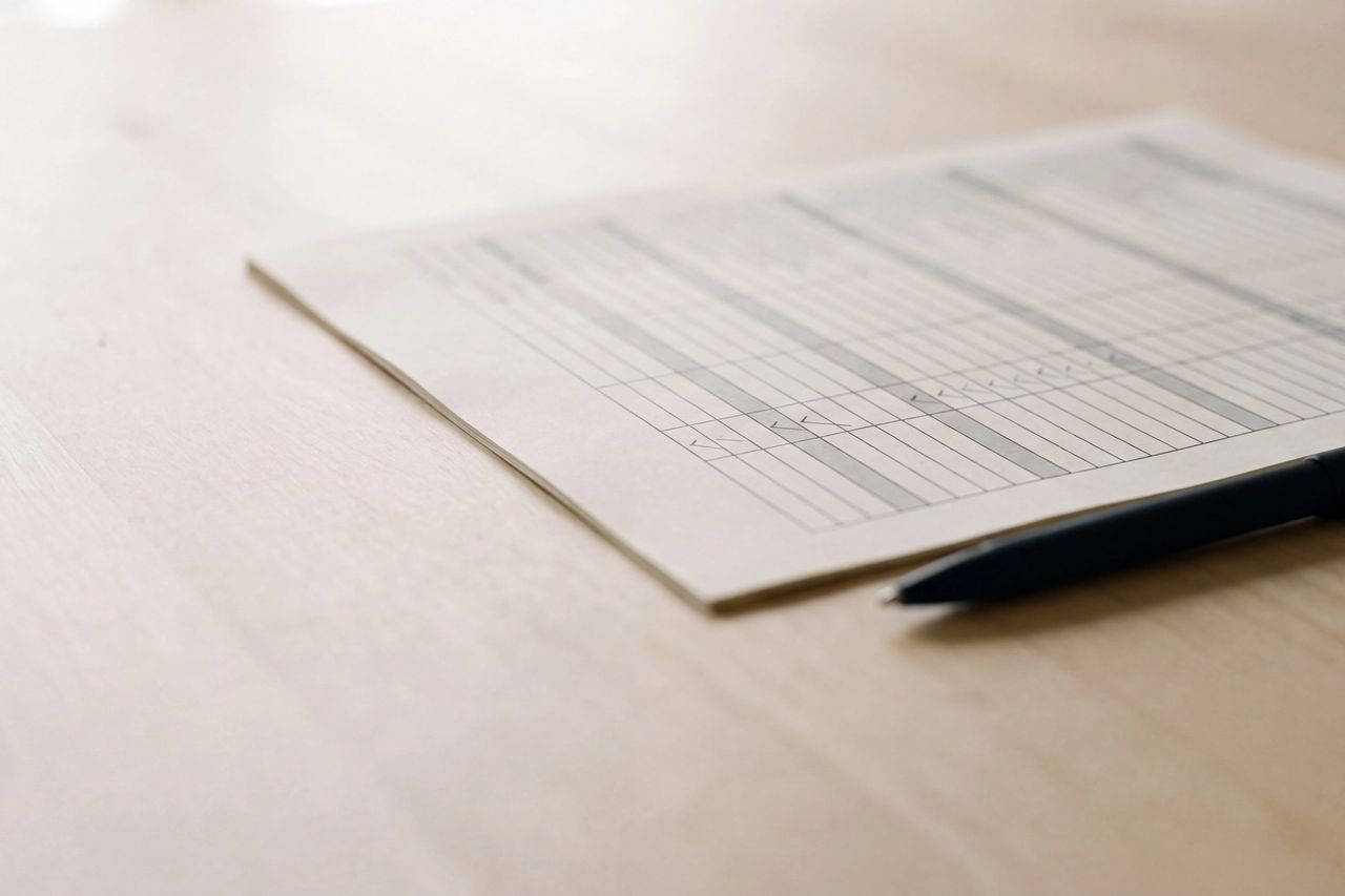 A printed control table with checkmarks lying on a pale wood desk beside a black pen.