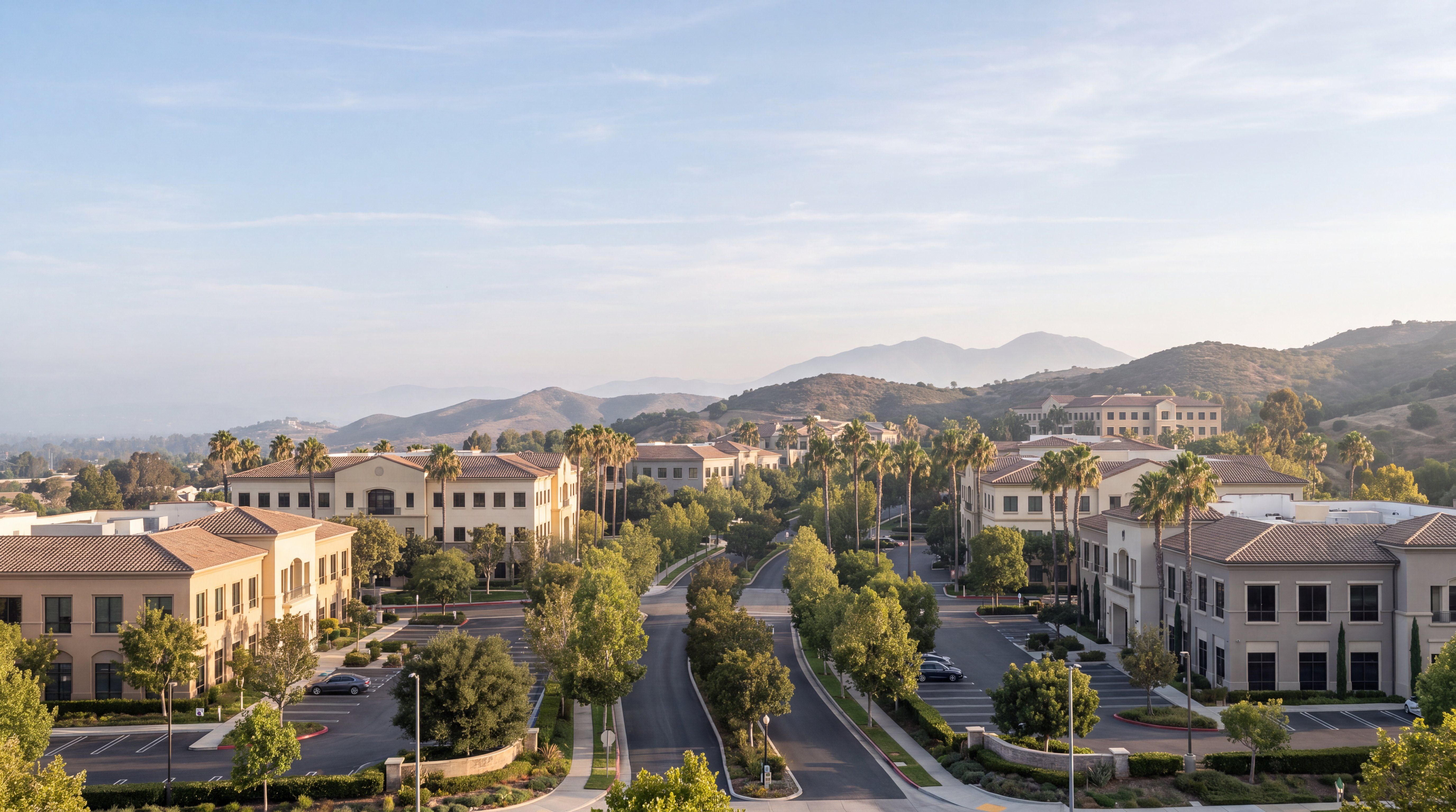 Low-rise office buildings with tile roofs among oaks and palms in the south Orange County foothills, with Saddleback Mountain in the morning haze beyond.