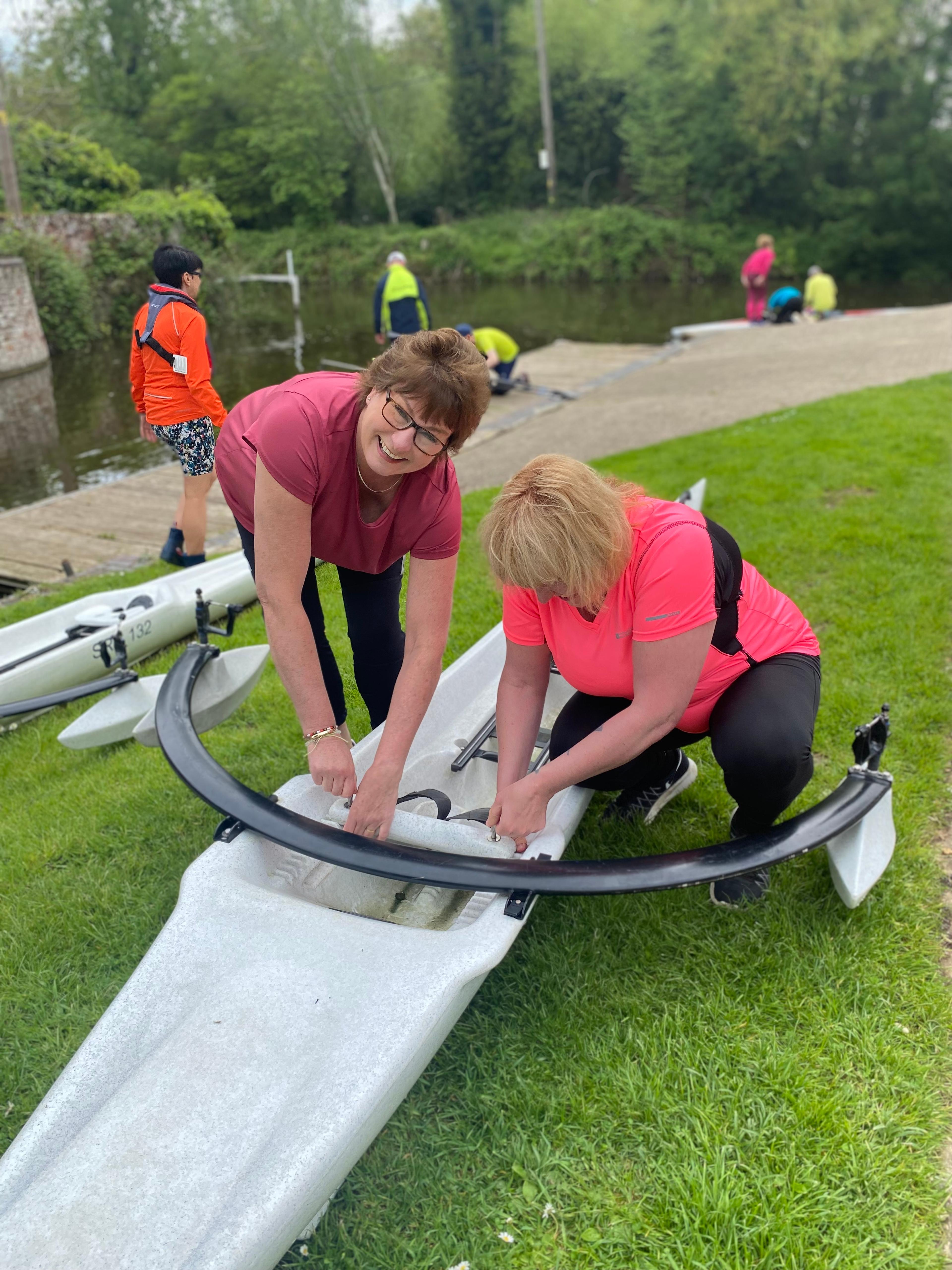 Two ladies adjust the footplate on a plastic training scull as it sits on the Spring green grass. 