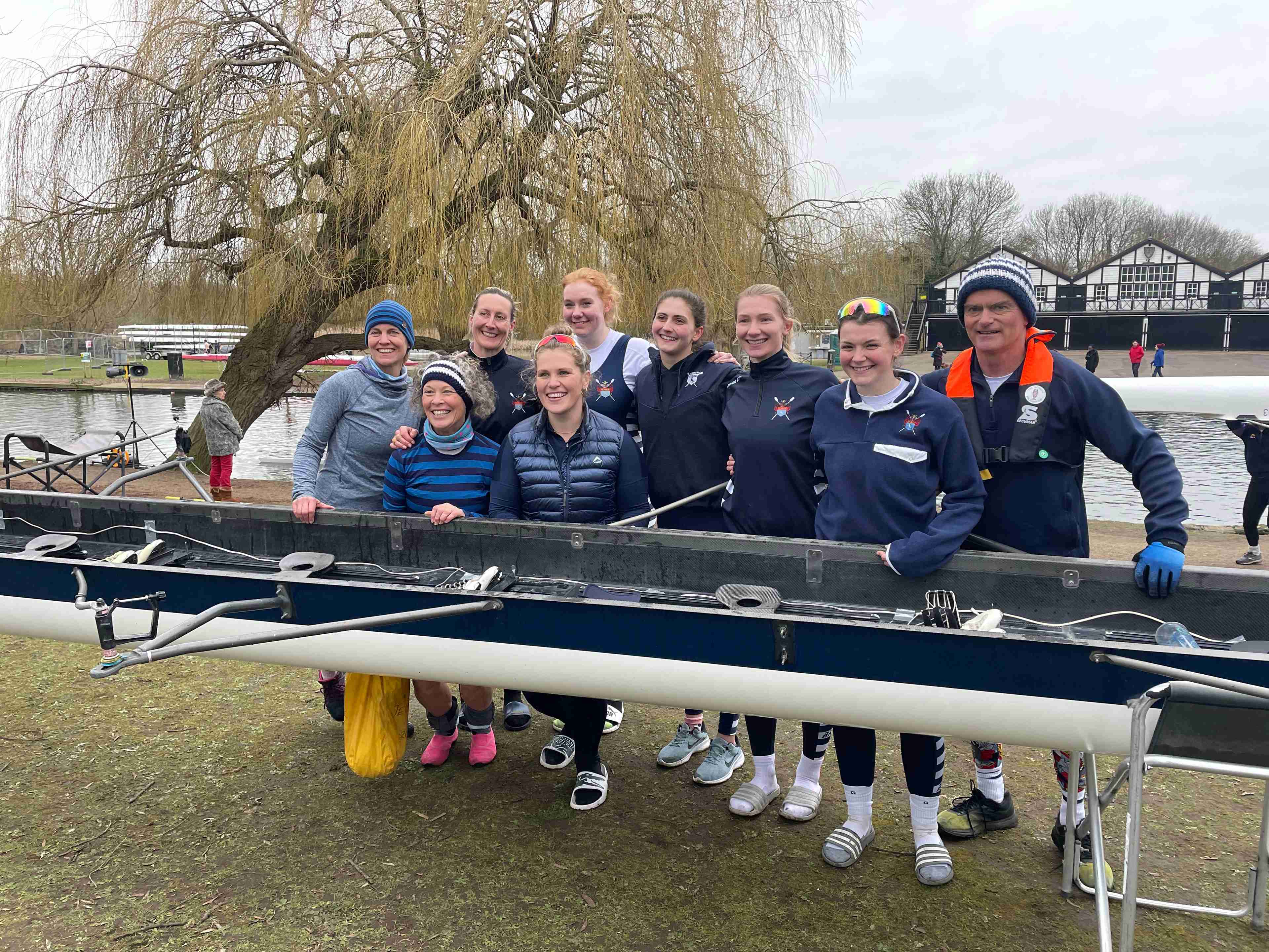 Sudbury’s victorious women’s eight stands behind their boat, which is on trestles on the foul-fouled patch of grass on the bank opposite Bedford Rowing Club. There's a weeping willow growing at a 45 degree angle in the background. 