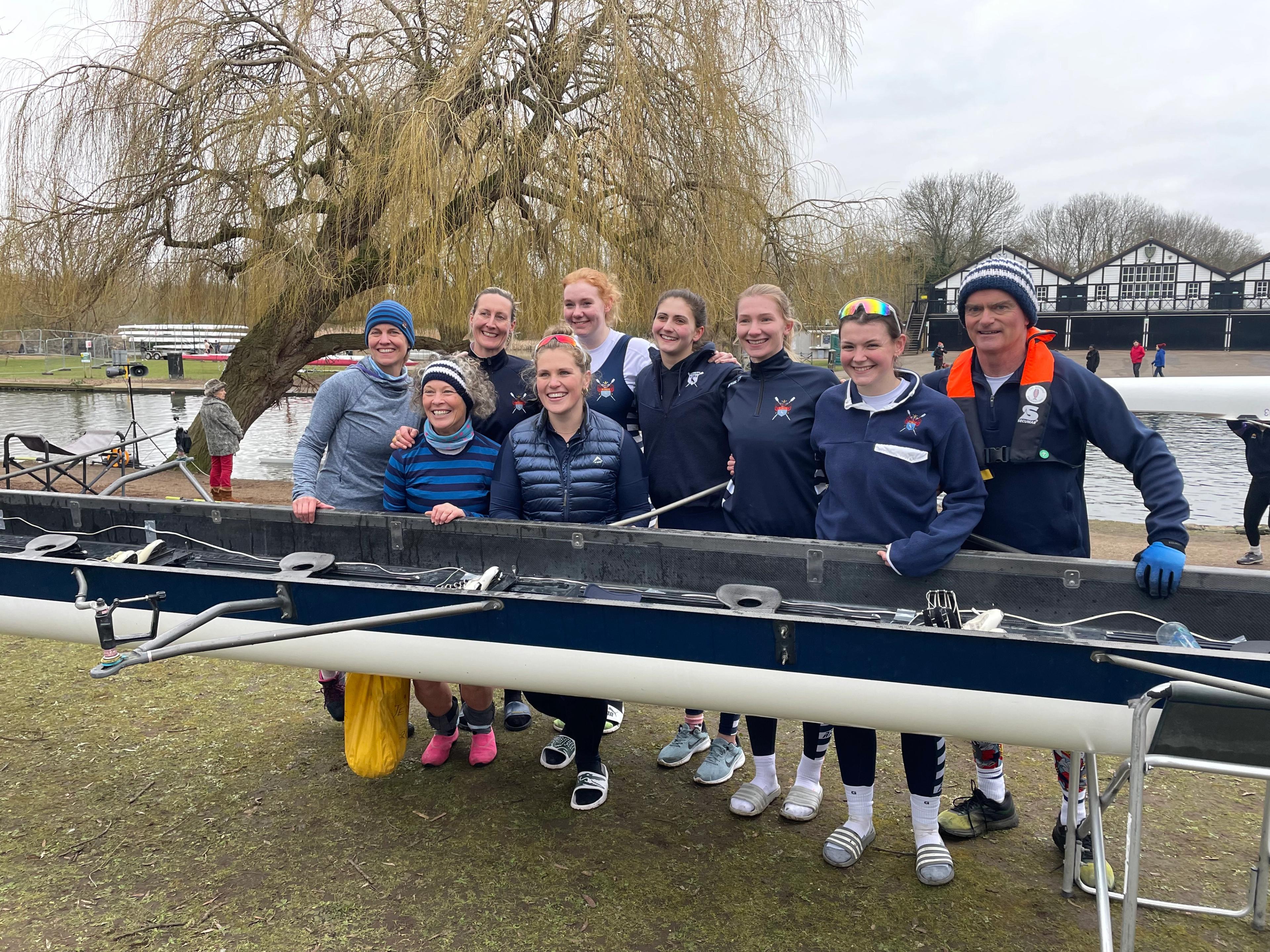 Sudbury’s victorious women’s eight stands behind their boat, which is on trestles on the foul-fouled patch of grass on the bank opposite Bedford Rowing Club. There's a weeping willow growing at a 45 degree angle in the background.