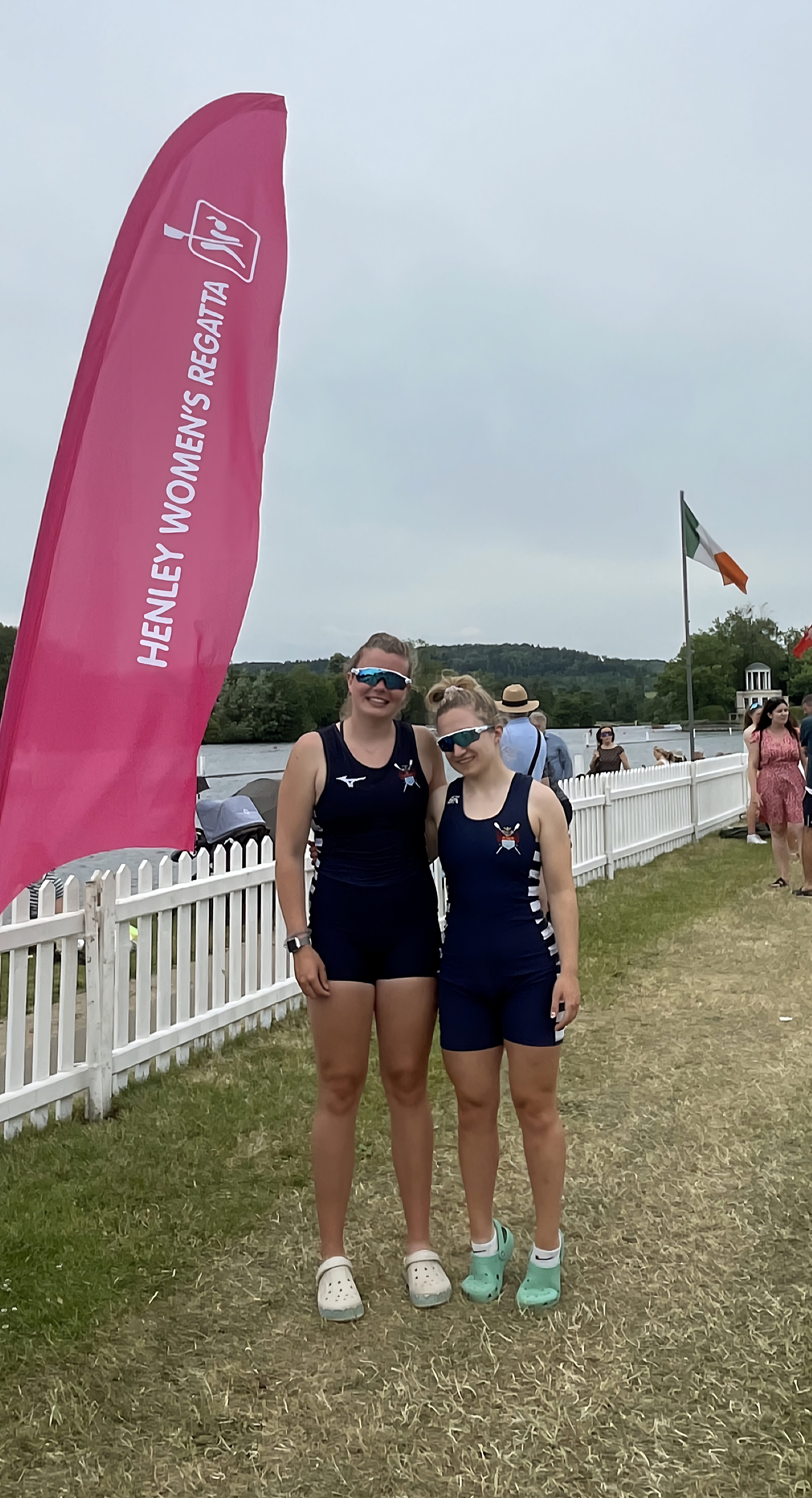 Martha Bullen and Amelia Moule from Sudbury Rowing Club standing together in front of a white picket fence, with the Henley Women's Regatta pink flag prominently displayed. They are wearing rowing kit and reflective sports sunglasses. 