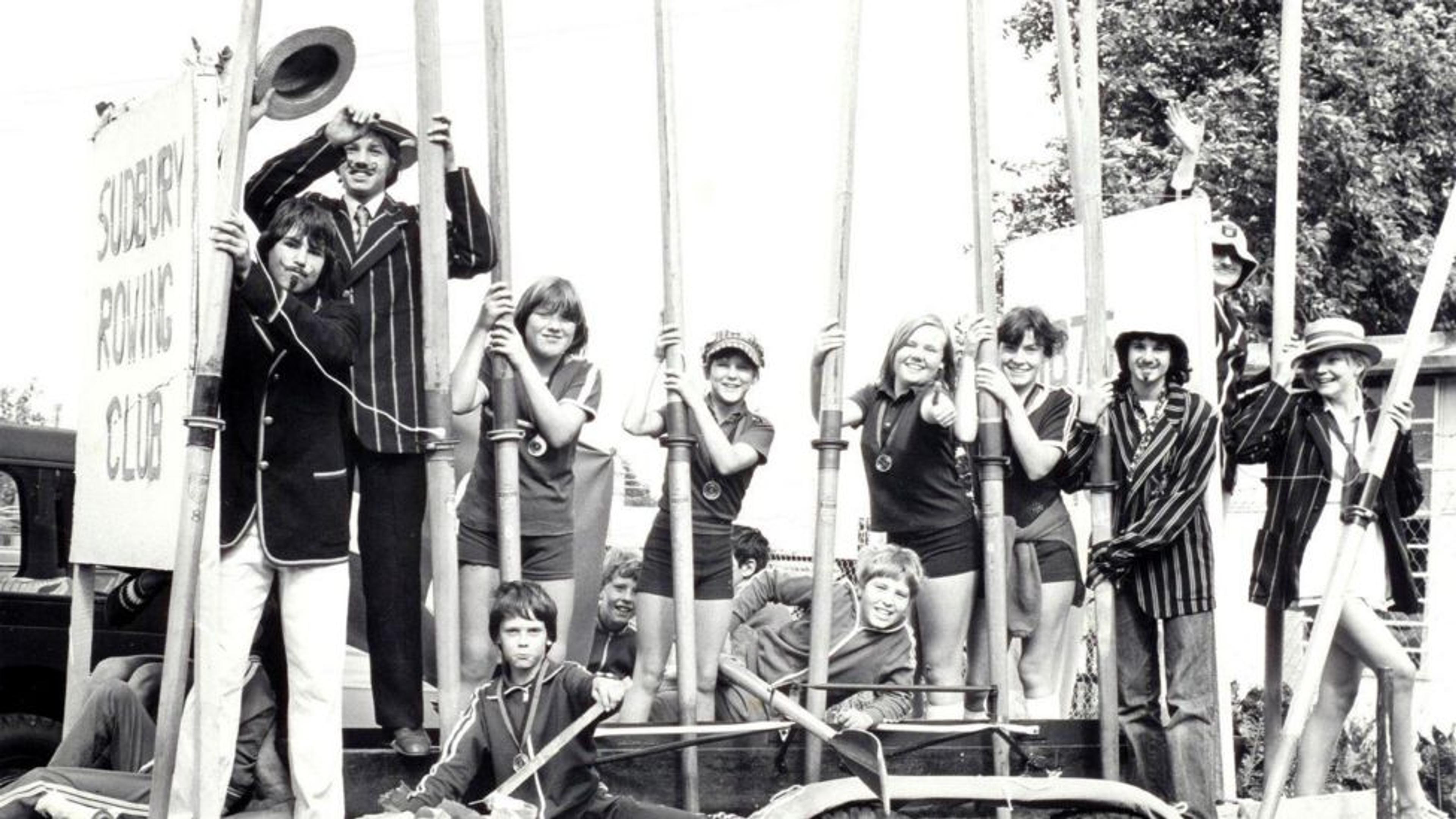 Black and white archival shot of a past club event. Male members wear blazers and support a large wooden sign stating the clubs name. Lady rowers in polo shirts hold up their blades while wearing medals. 
