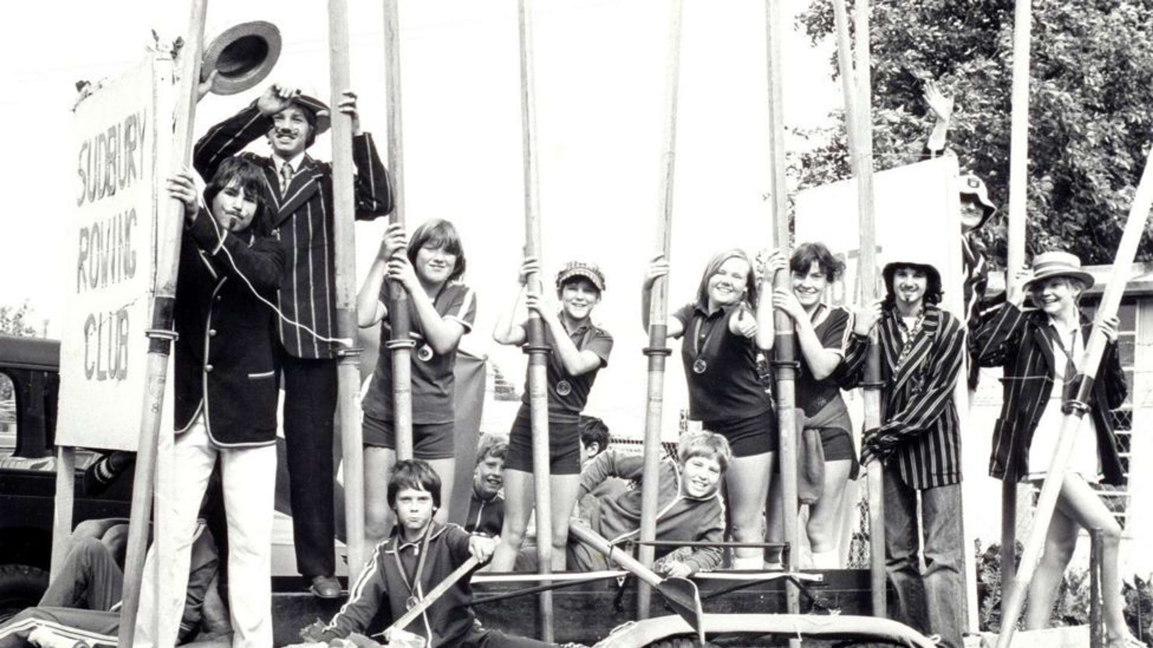 Black and white archival shot of a past club event. Male members wear blazers and support a large wooden sign stating the clubs name. Lady rowers in polo shirts hold up their blades while wearing medals.