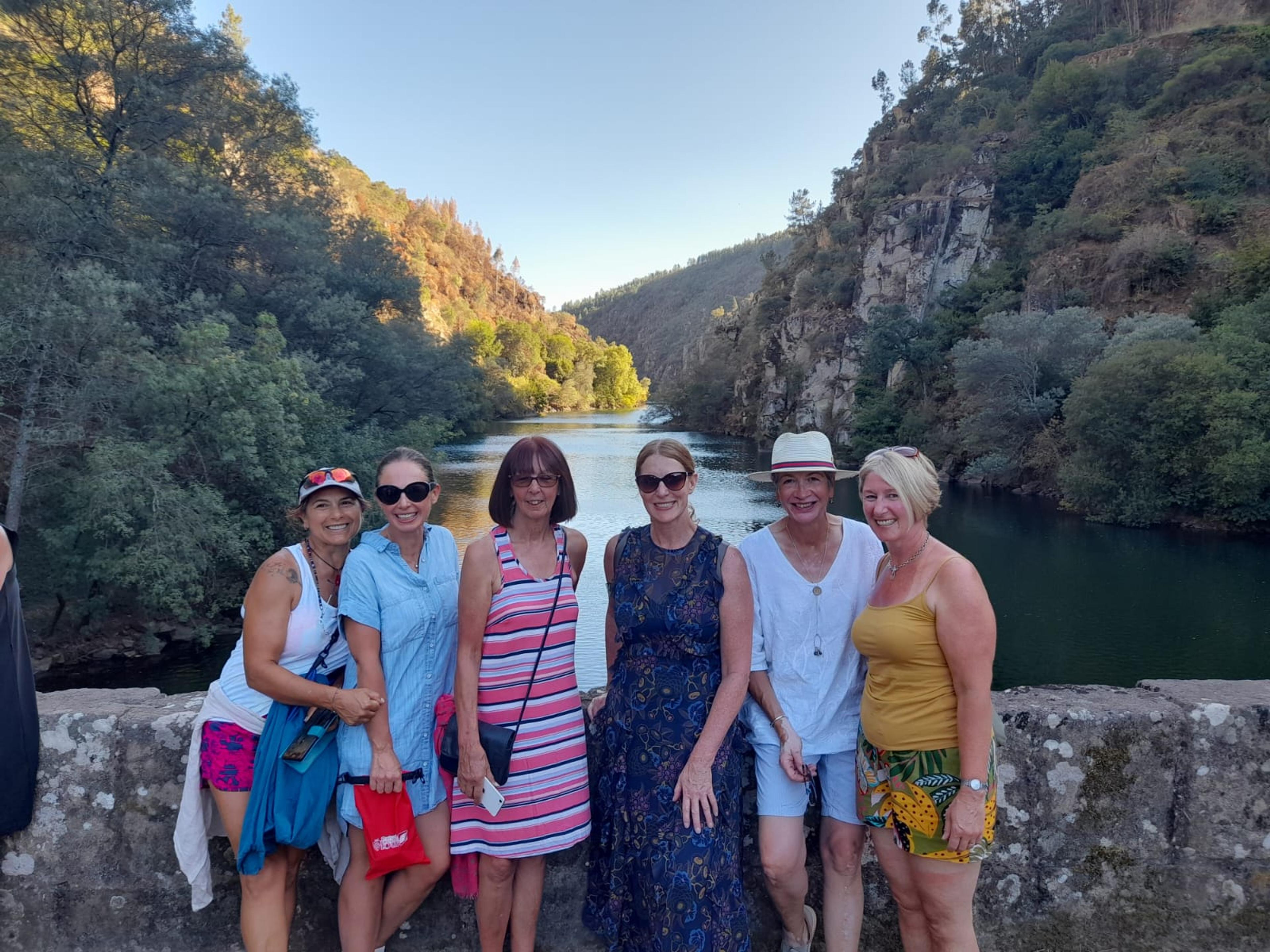 The seven Sudbury rowers pose for a group photo on a lichen-covered bridge over a river, at the bottom of a steep river valley, whose sides a partly rocky but mostly covered with trees that look well adapted to fairly dry conditions. 