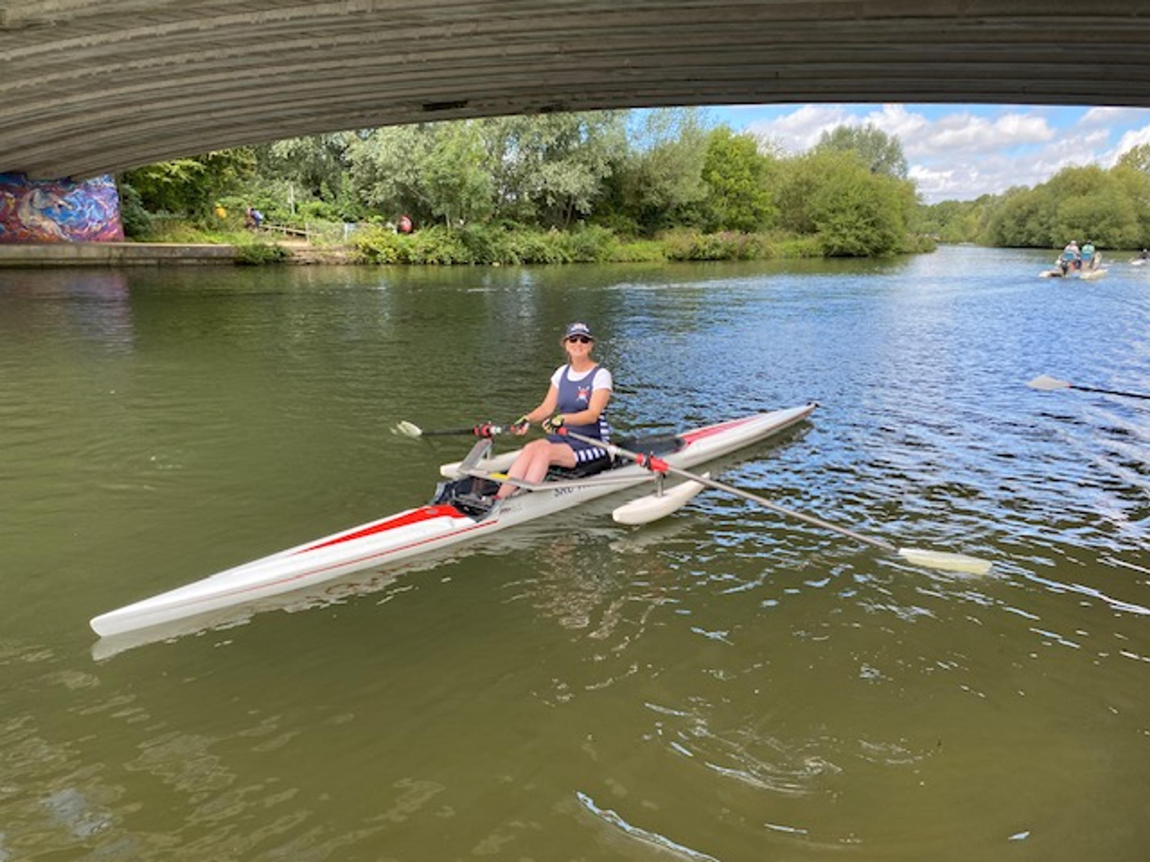 An adaptive single sculler under a bridge on the opaque green water of the Thames in Oxford. 